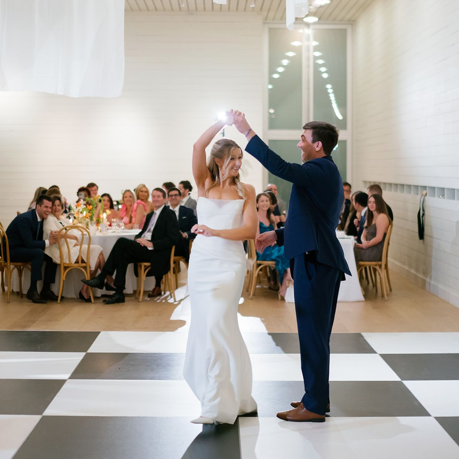 A couple dances on a black-and-white checkered dance floor in a bright venue while guests watch from tables.