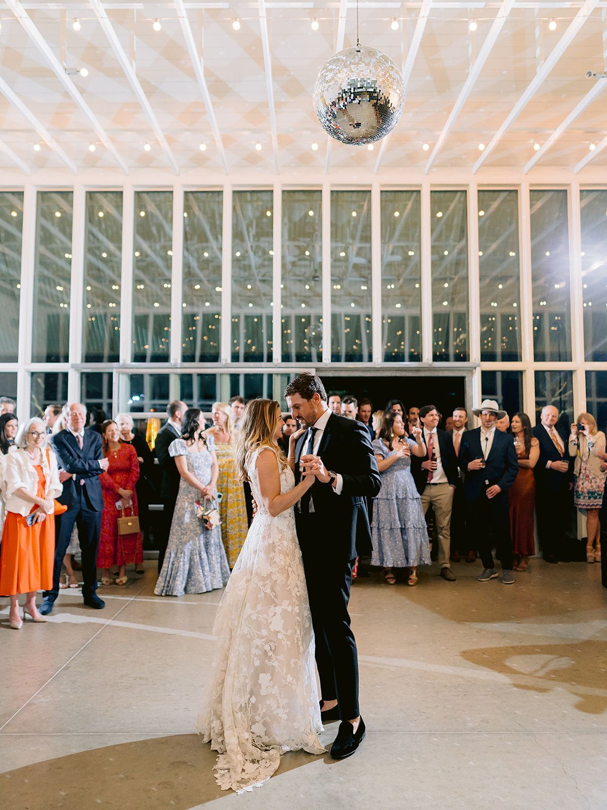 A couple dances at their wedding reception in Prospect House's Gallery with a glass wall and a disco ball, surrounded by guests.