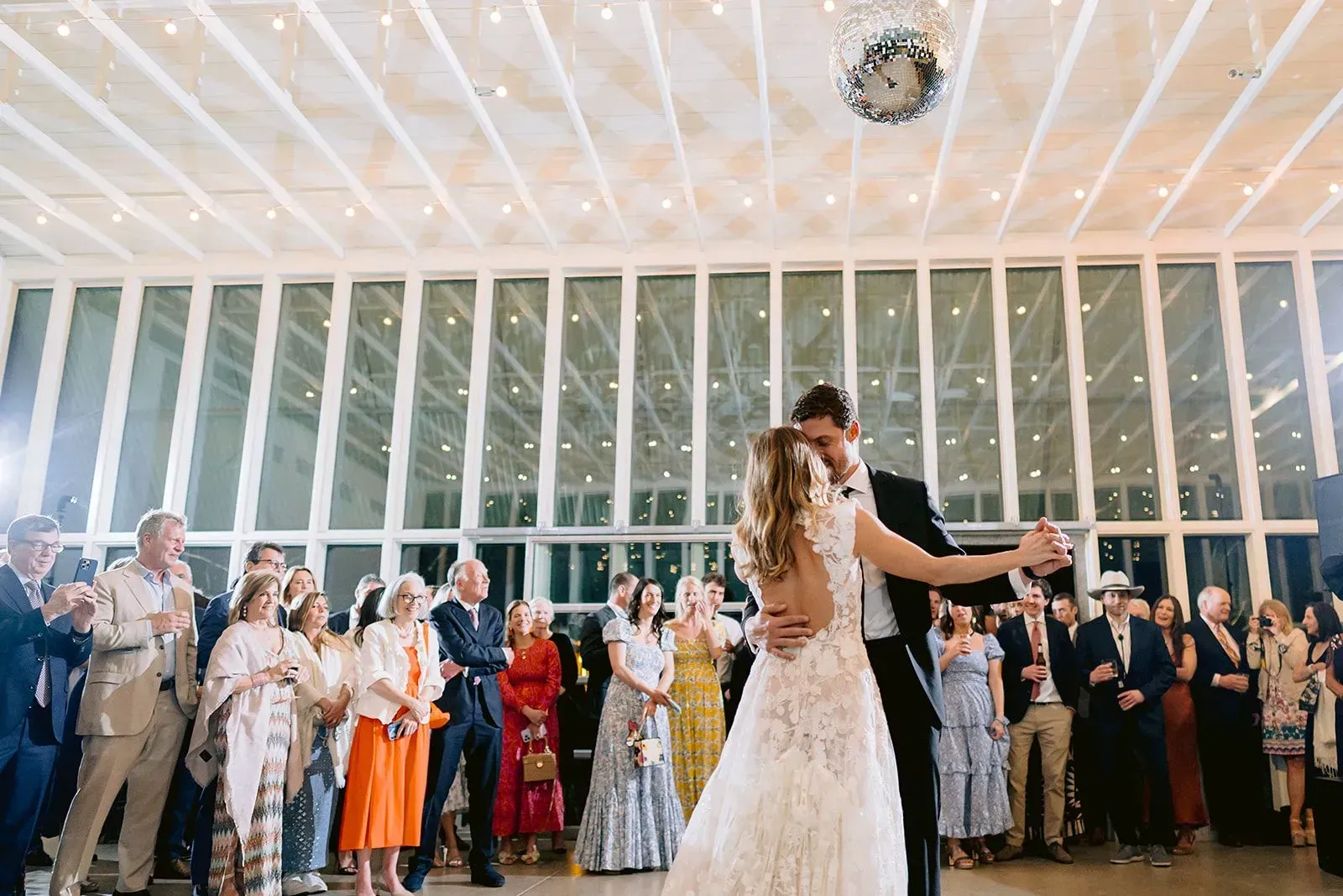 A couple dances during their wedding reception in a large, bright hall with floor-to-ceiling windows and guests watching.