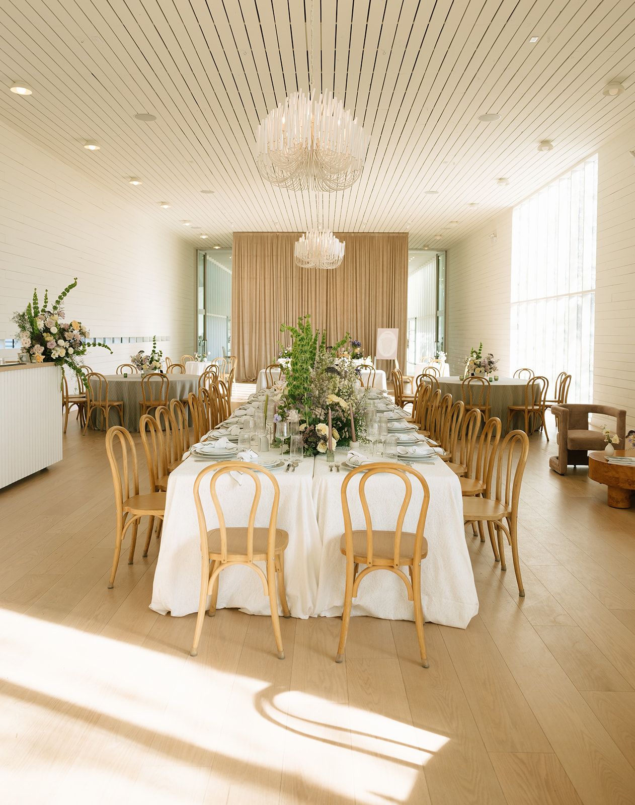 A long, elegant dining table with white linens and floral centerpieces in a bright, modern hall with wood floors.