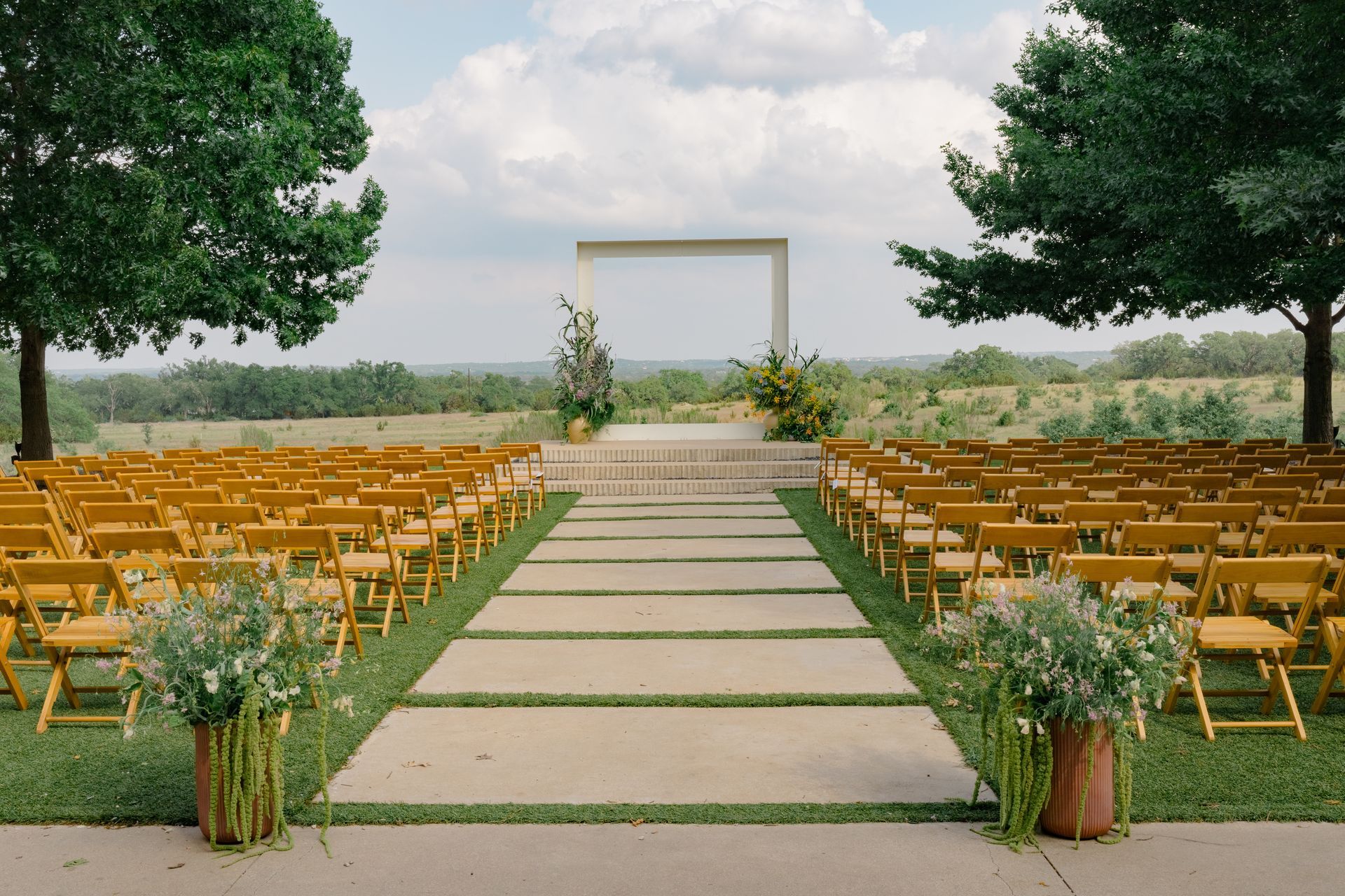 An outdoor wedding aisle lined with rows of wooden chairs leads to a minimalist arch overlooking a scenic, grassy field.