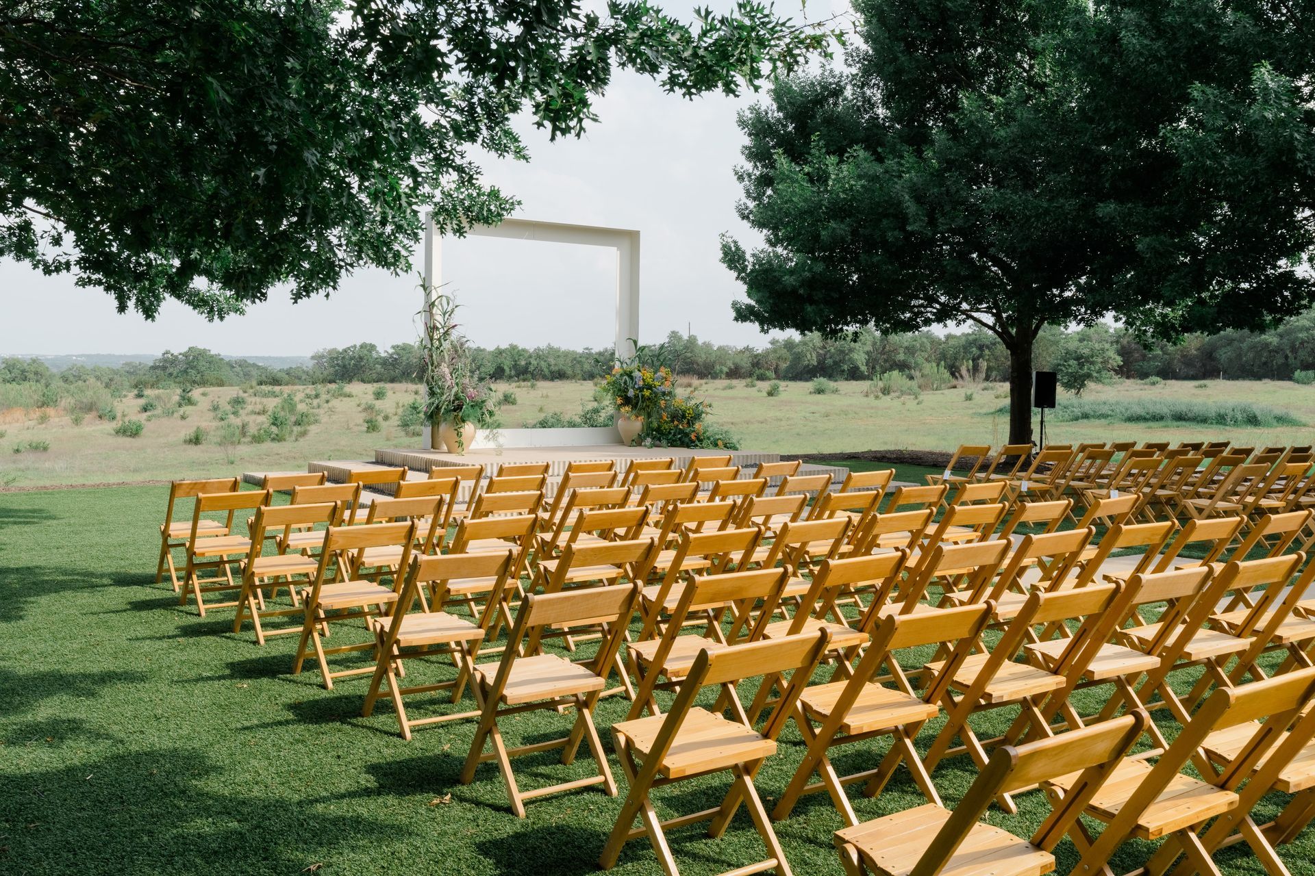 An outdoor wedding venue In the Texas Hill Country with rows of wooden chairs facing a modern rectangular arch on a grassy lawn under trees.