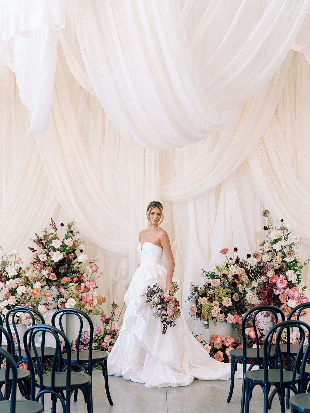 A person in a white wedding gown stands between two floral arrangements in front of a white draped backdrop with chairs designed by Native Bloom.