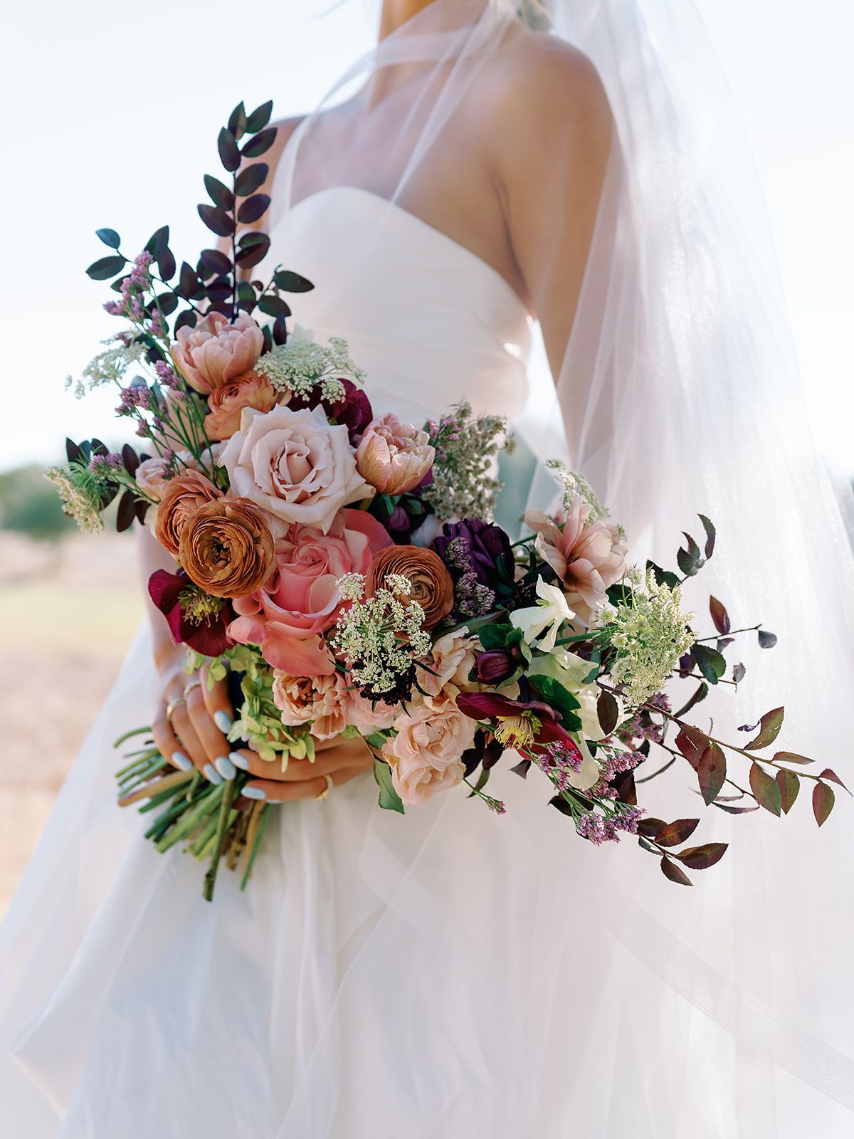 A bride in a white wedding dress holds a bouquet of roses, dark foliage, and wildflowers in earthy, warm tones designed by Native Bloom Floral. 