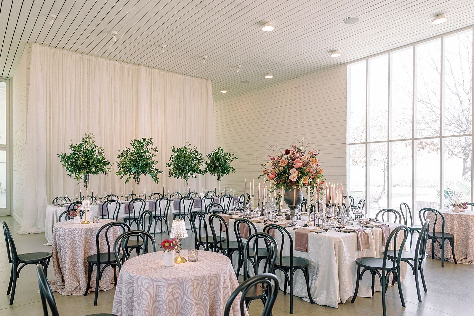 Elegant event space with round tables, draped linens, floral centerpieces, and black chairs beneath a slatted ceiling.