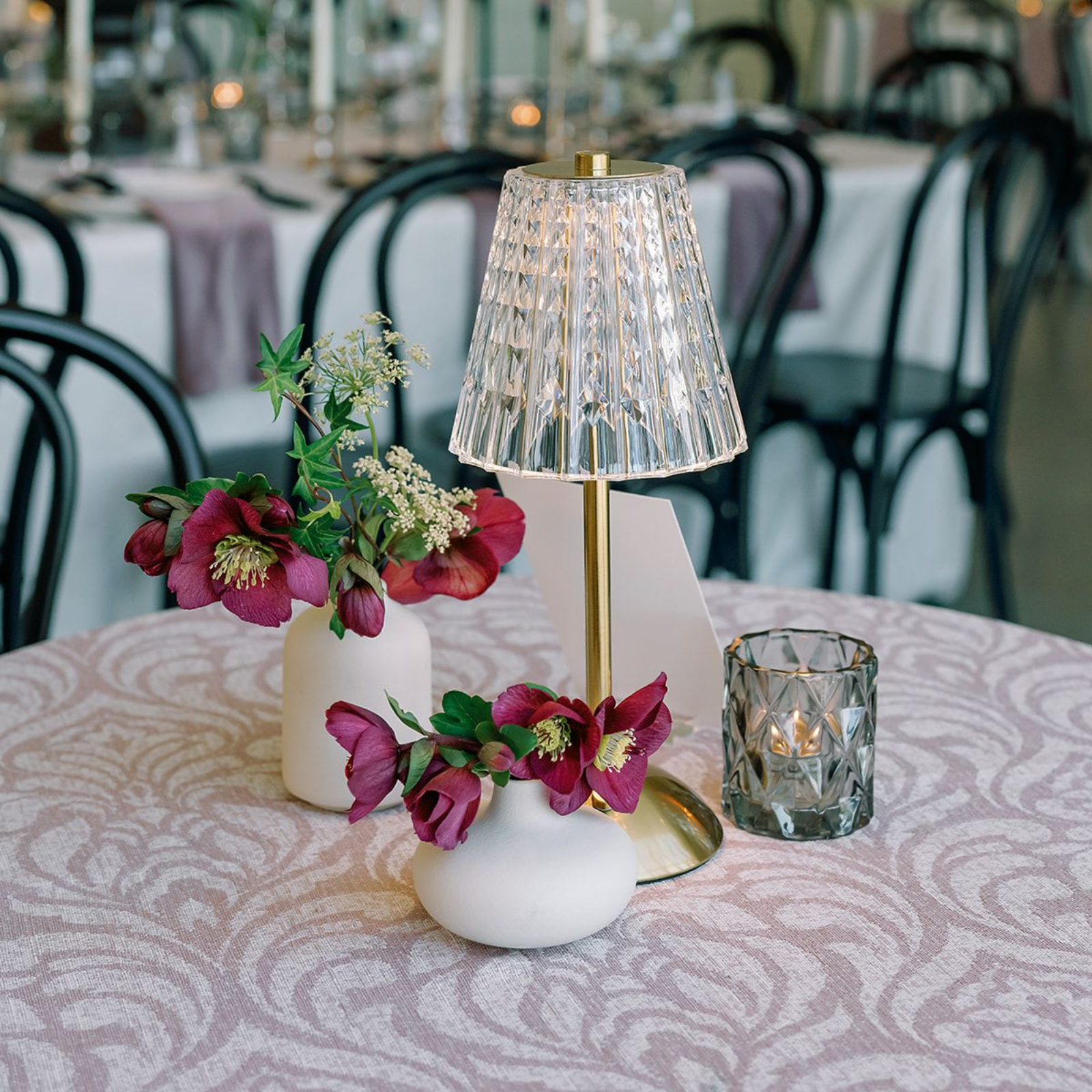A wedding table centerpiece with a gold cordless lamp, two white vases of dark pink hellebore flowers, and a glass candle.