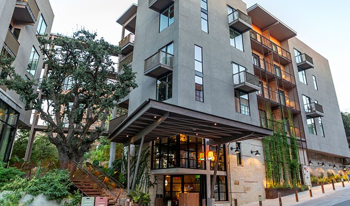 A large concrete building with patios off each window and natural tree at the entrance of it. 