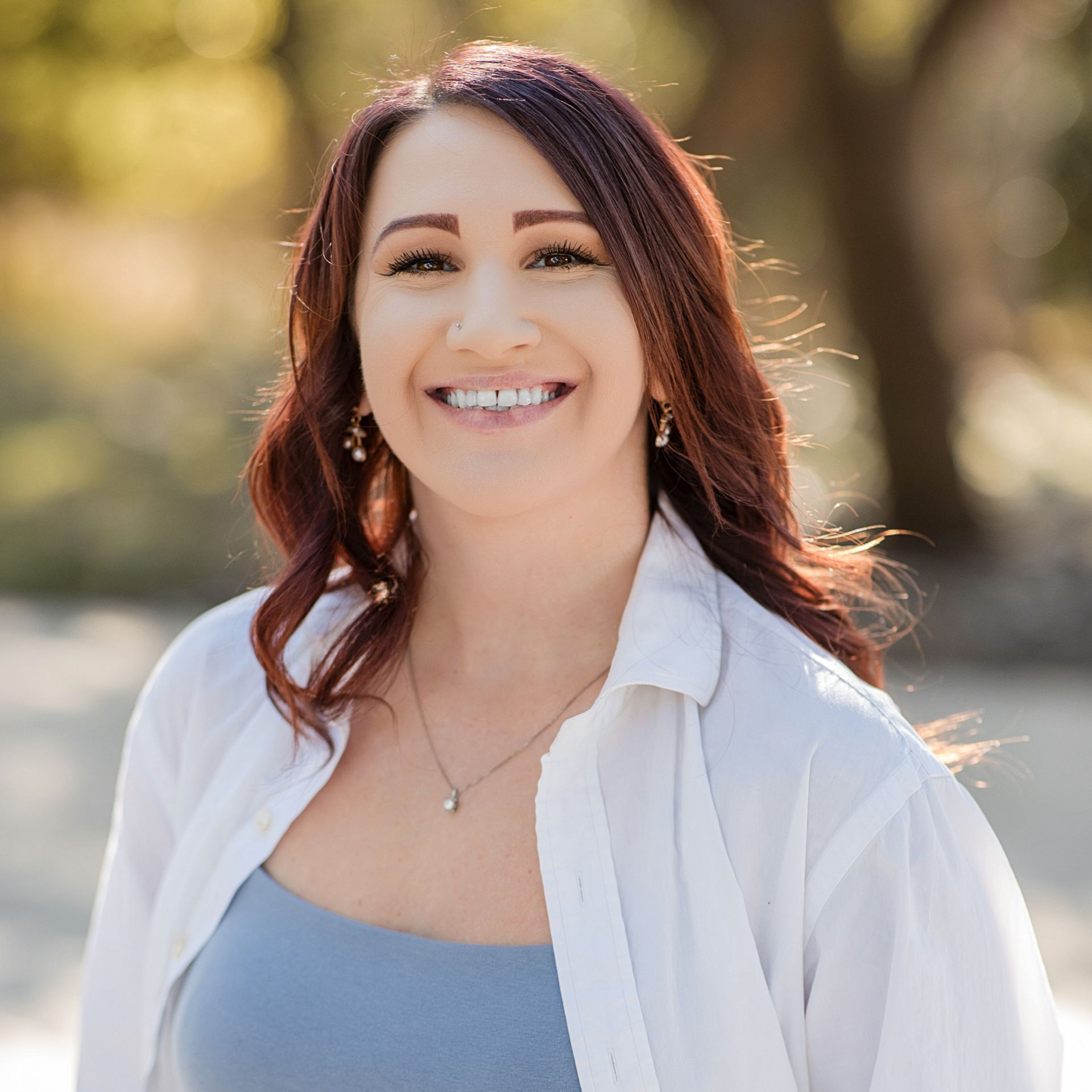 A smiling person with auburn hair wearing a white shirt and blue top, posing outdoors with a soft, blurred background.