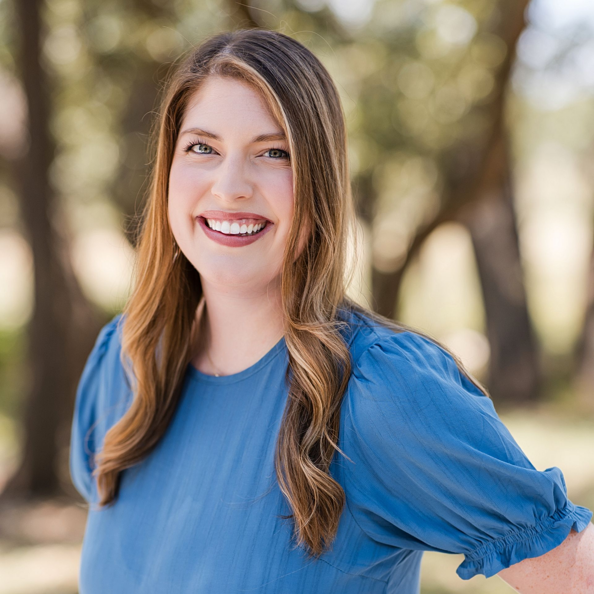 A smiling person with long light brown hair wearing a blue, short-sleeved top, posed outdoors against a blurred background.