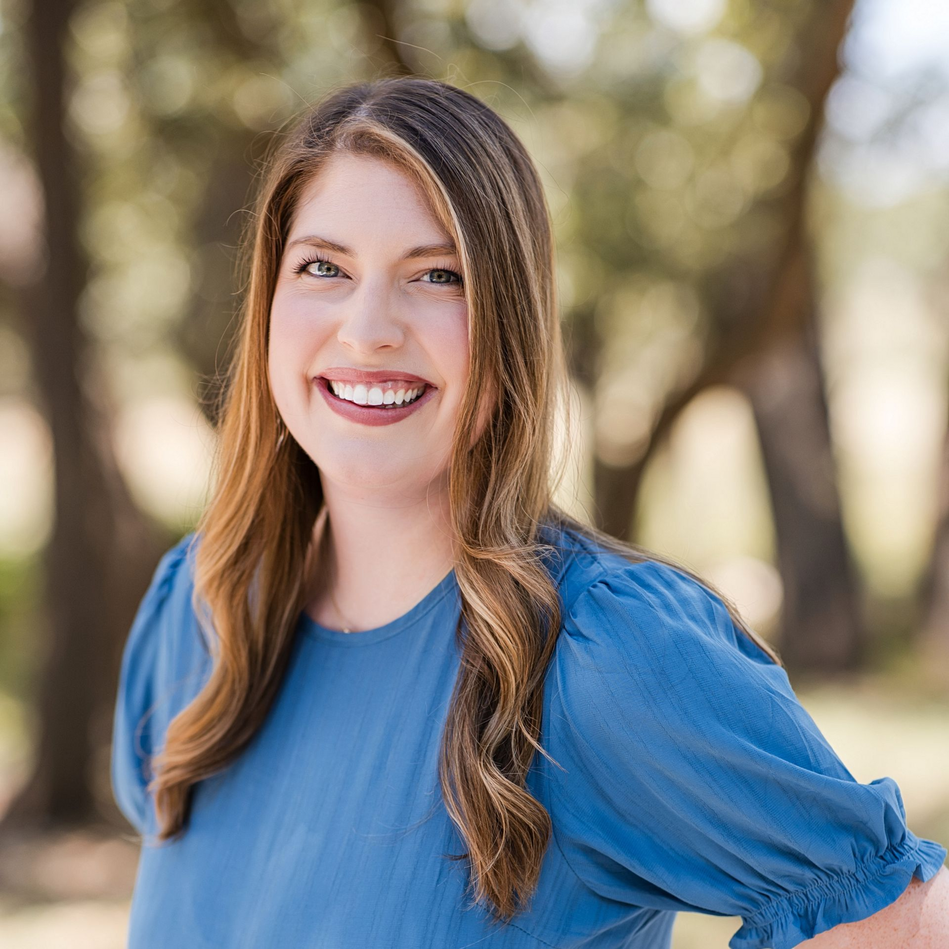 A person with shoulder-length brown hair wearing a blue shirt smiles warmly in an outdoor, wooded setting.