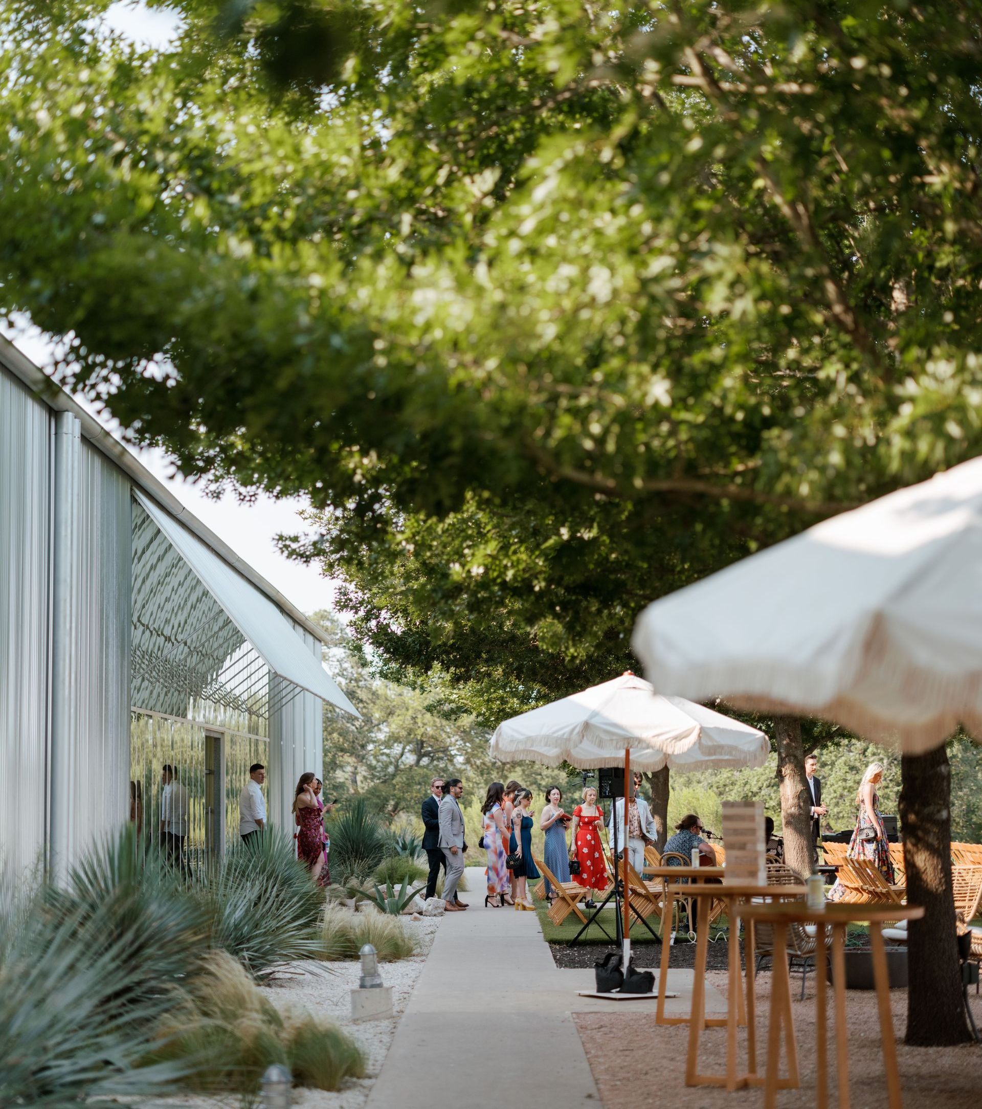 Outdoor wedding venue with a paved path, white patio umbrellas, and guests mingling near a white structure under trees.
