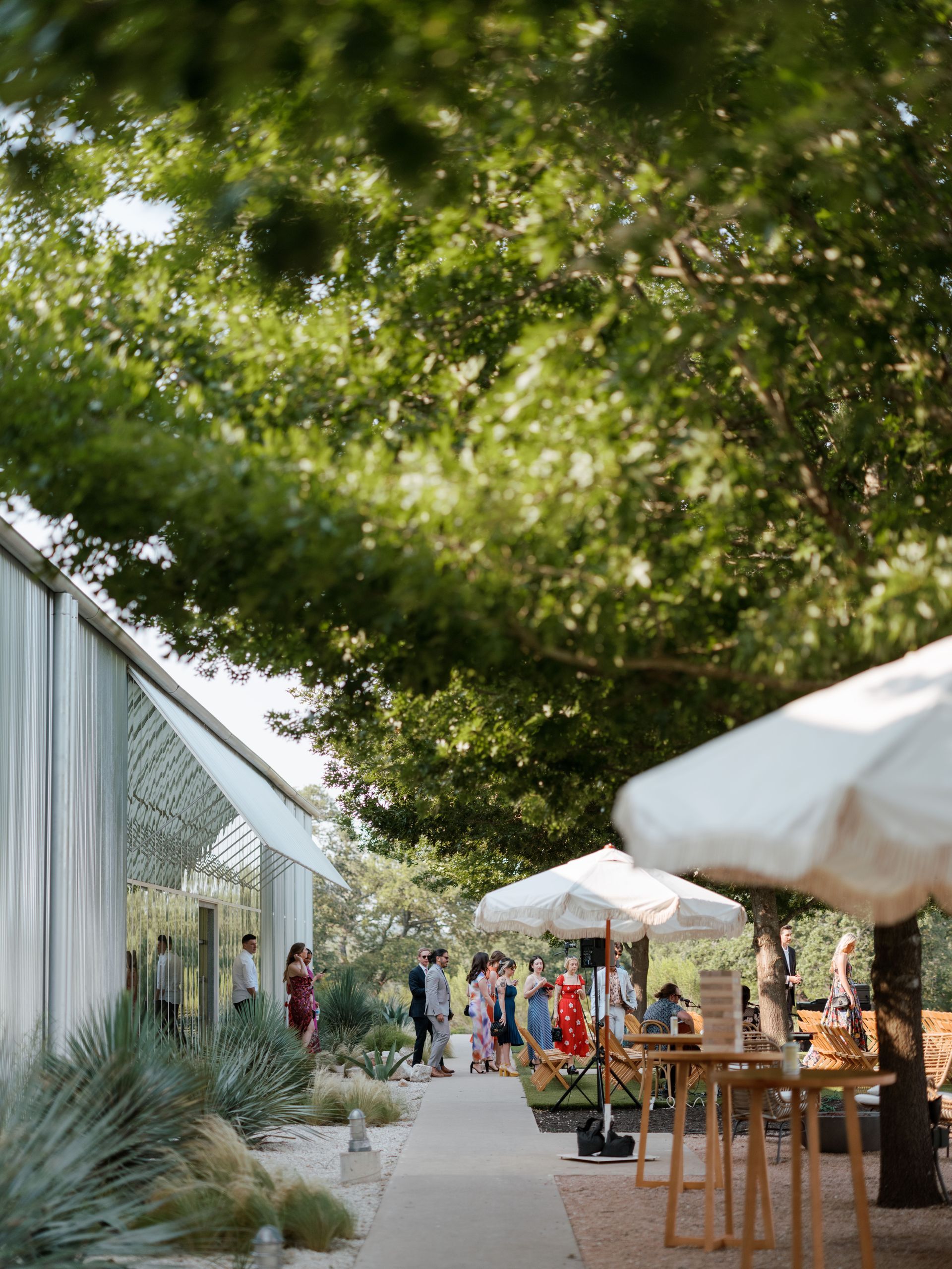 Guests gather at an outdoor reception under trees and white umbrellas next to a modern tent structure.