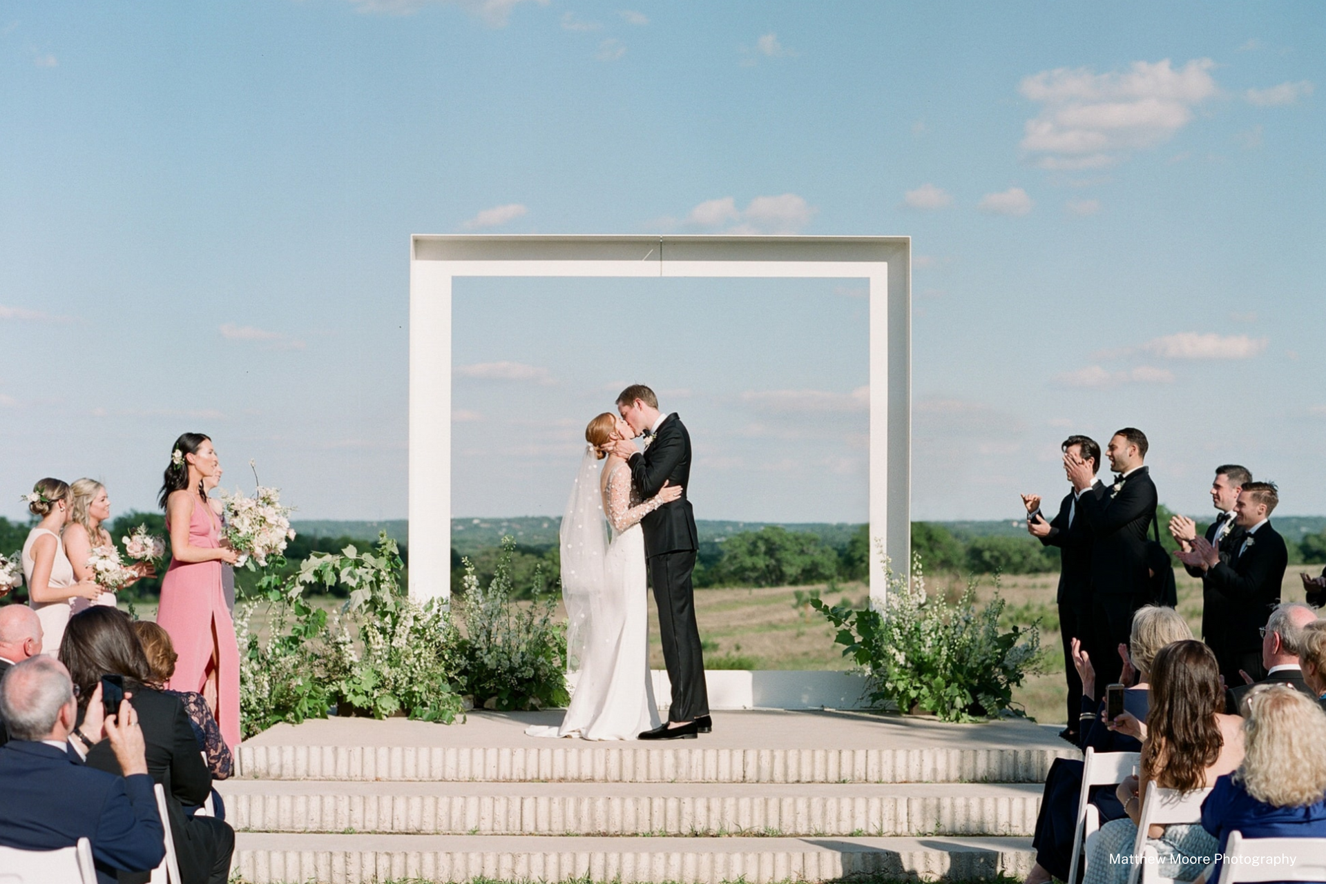 A couple kisses on a raised outdoor stage framed by a white arch, with wedding party members standing on either side.