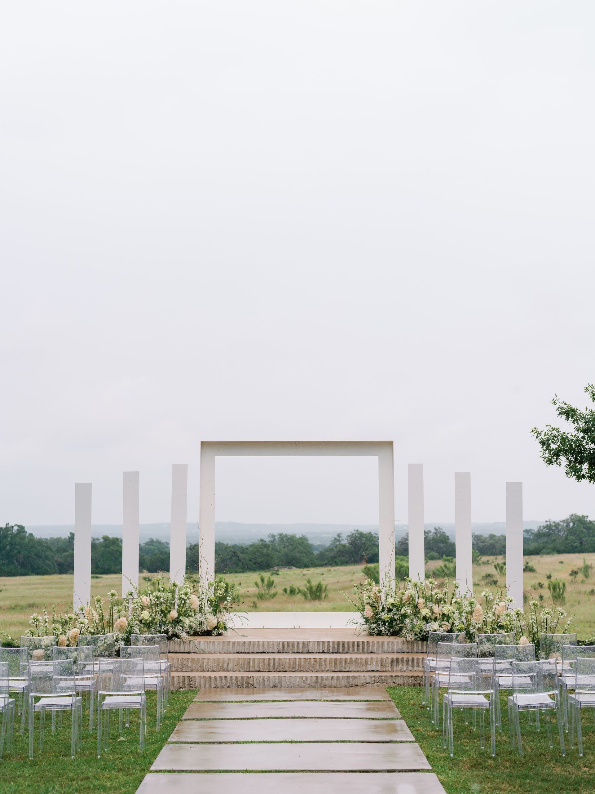An outdoor wedding altar with a white frame and pillars on a grassy hill, leading to a concrete path and rows of chairs.