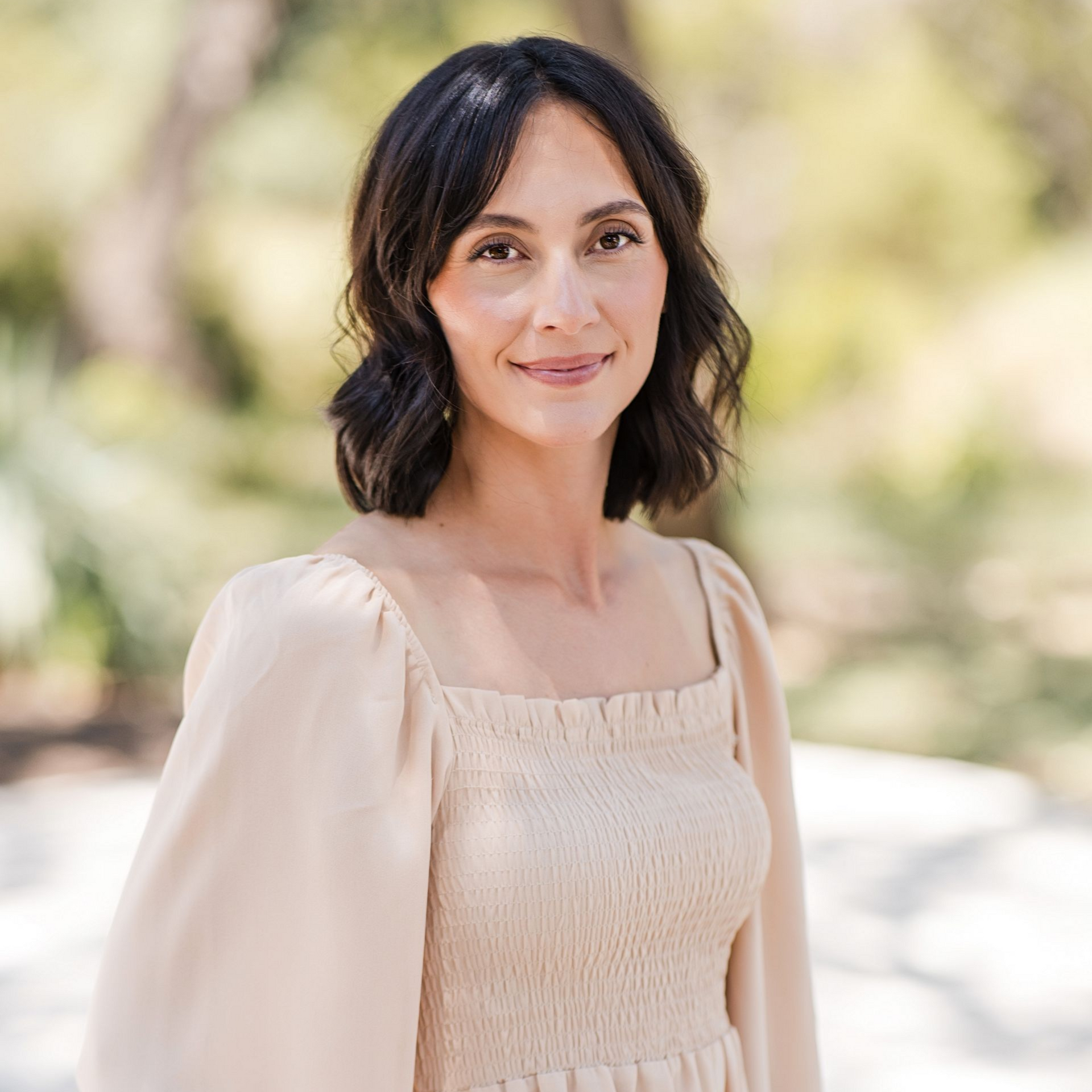 A person with dark hair wearing a light beige smocked-top dress, standing outdoors with a soft, blurred natural background.