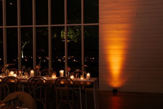 A candlelit dinner table set before large windows at night, with an amber uplight casting a glow on a wooden wall.
