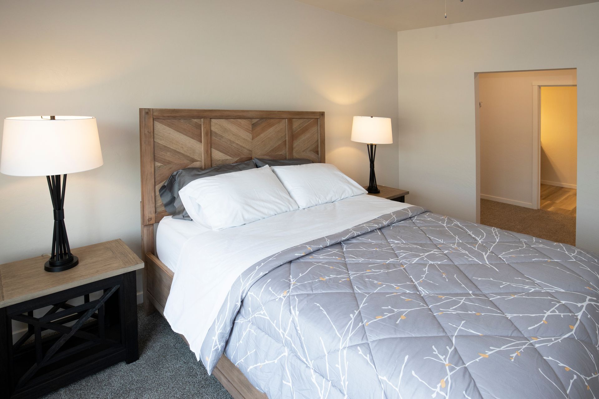 Bedroom with a wooden headboard, gray bedding, two lamps, and an open doorway to a hall.
