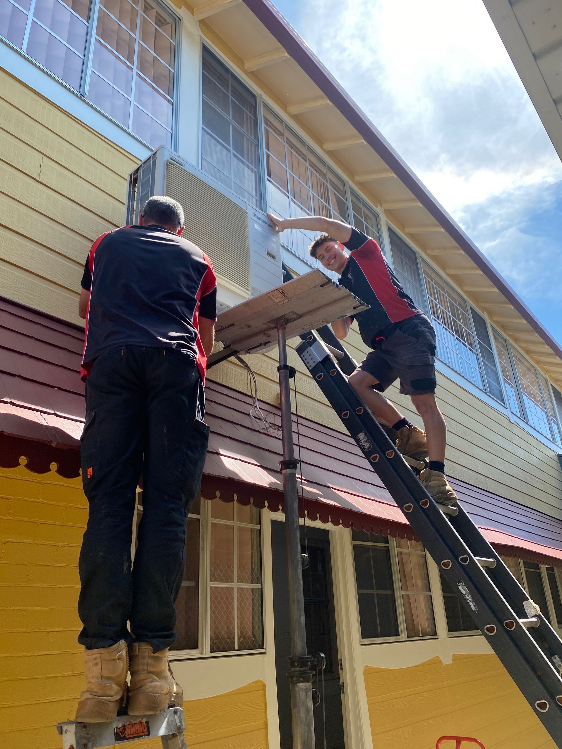 Two workers install an air conditioning unit on a building. One on a ladder, the other on scaffoldingairCoolman Airconditioning in Broken Head, NSW