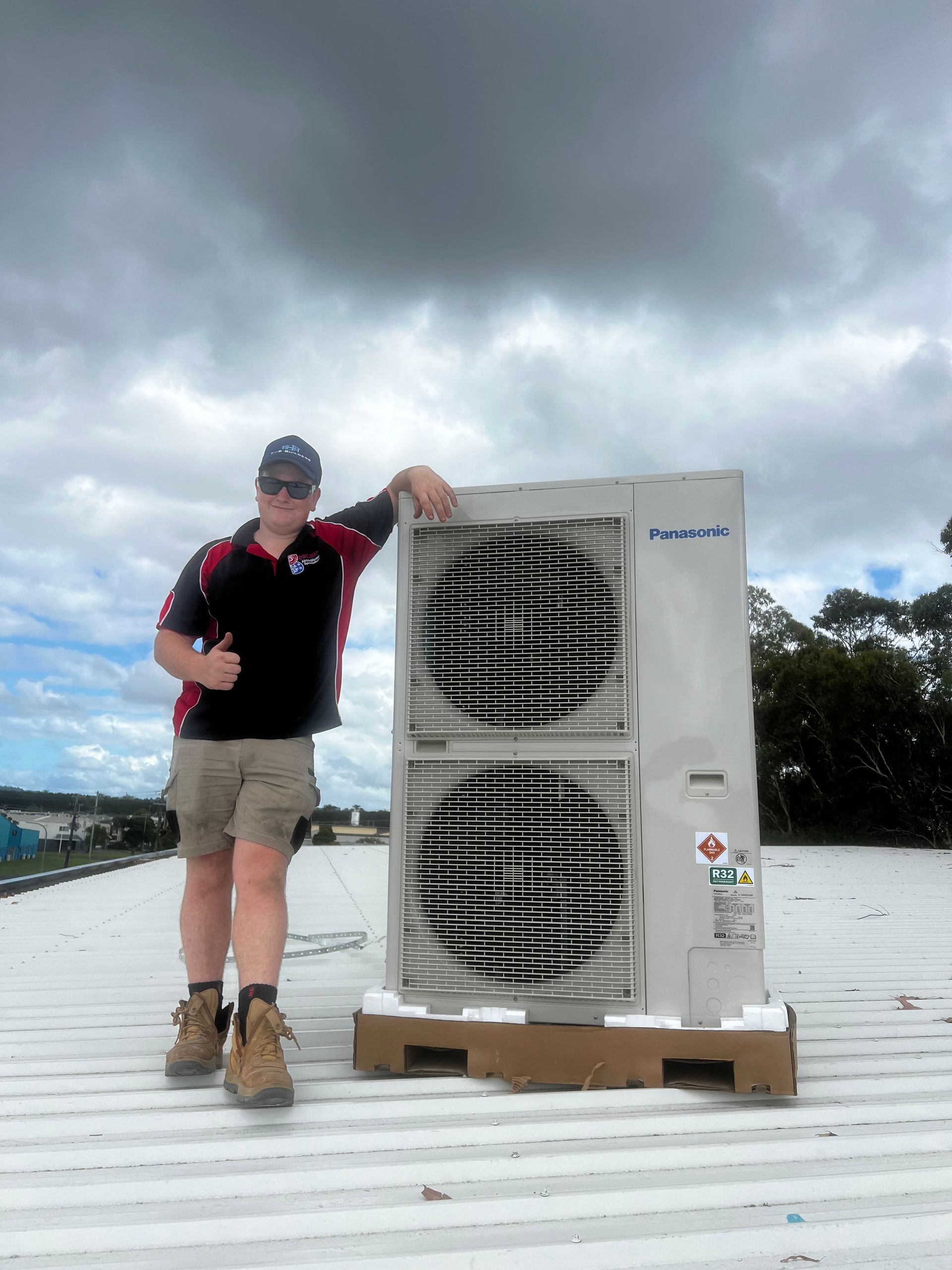 Man in work clothes thumbs up beside a large air conditioning unit on a rooftop. Cloudy sky - Coolman Airconditioning in Broken Head, NSW