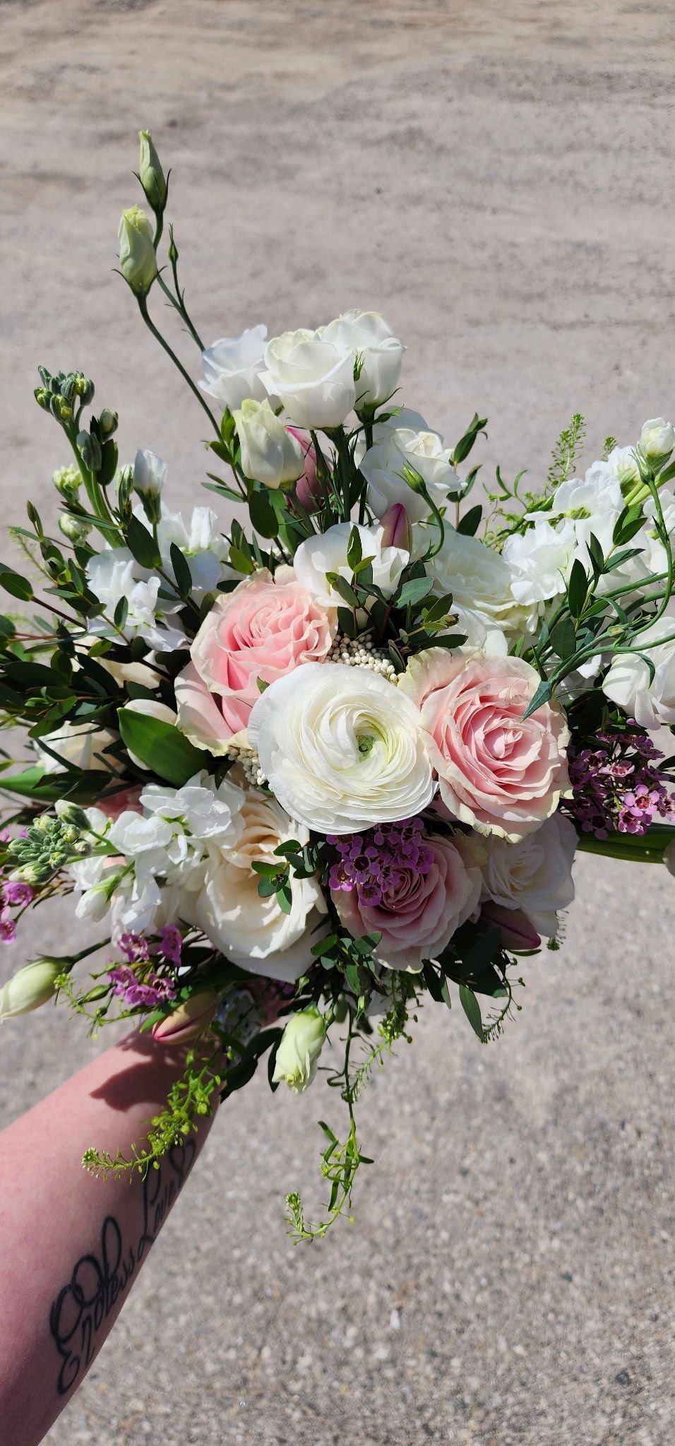 Handheld bouquet of white, pink, and lilac flowers with greenery against a concrete background