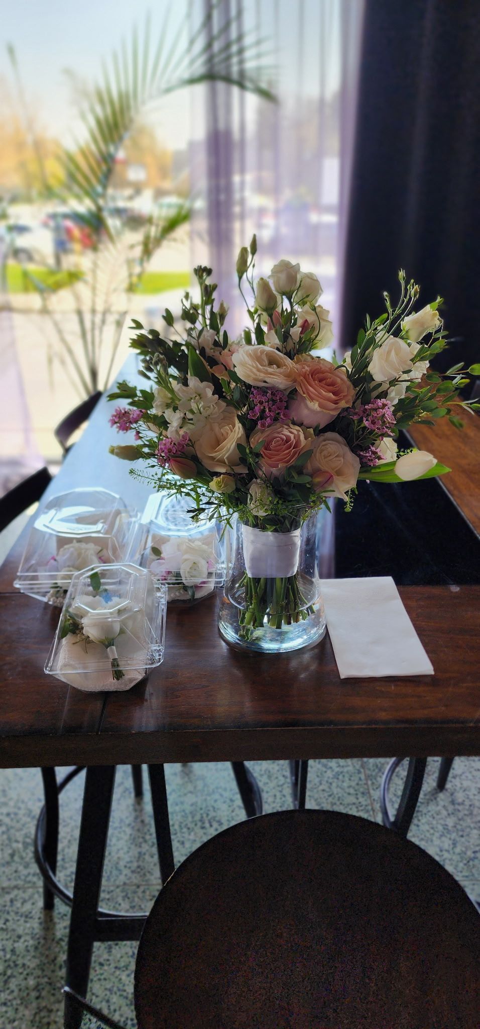 Flower arrangement on a wooden table with candles and a folded note, near a bright window.