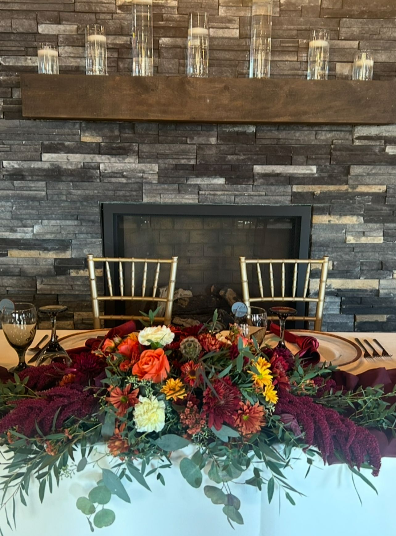 Flower arrangement on a table in front of a stone fireplace with two gold chairs and candles on the mantel