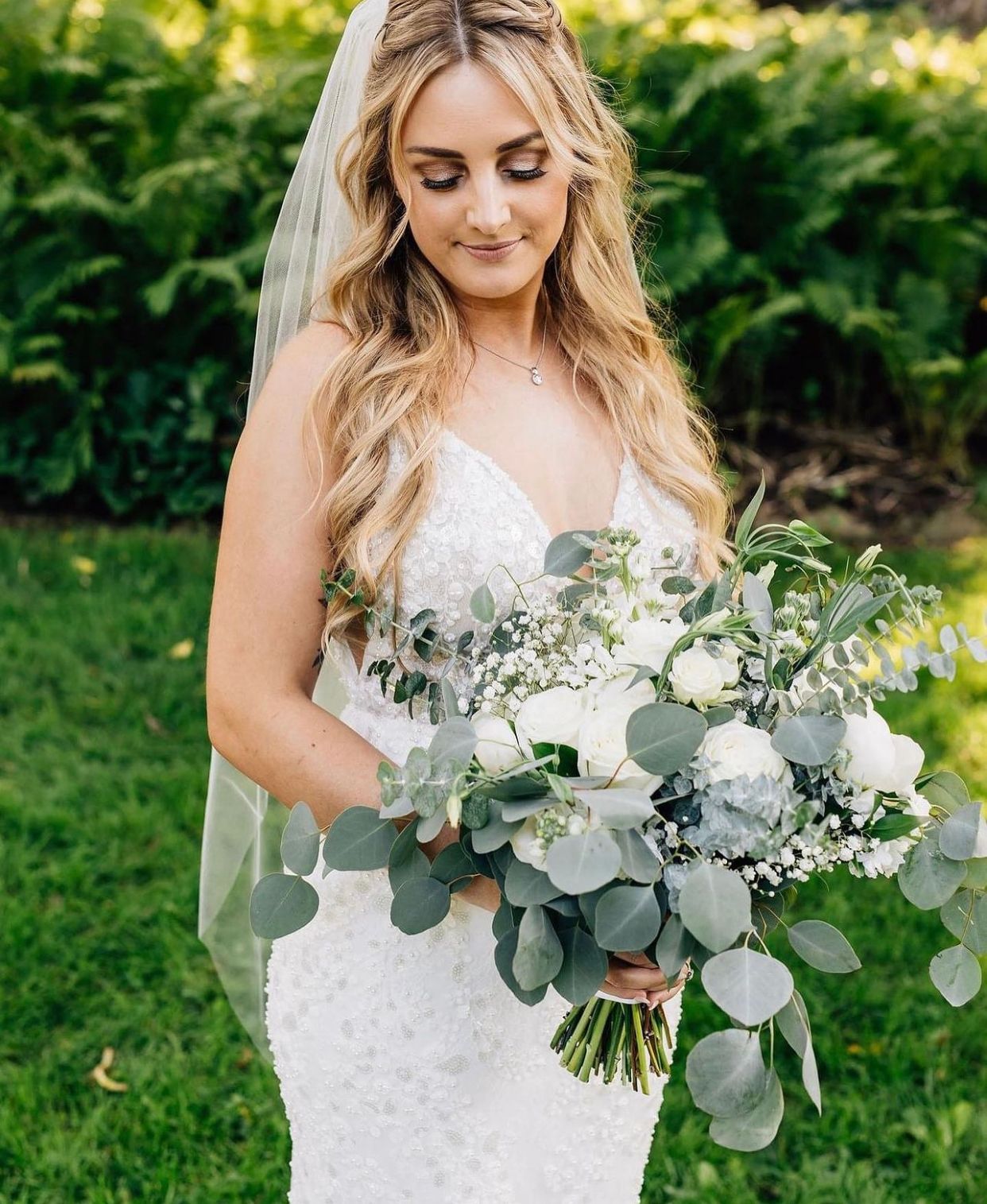 Bride in a white lace gown holding a large bouquet of eucalyptus and white flowers in a garden
