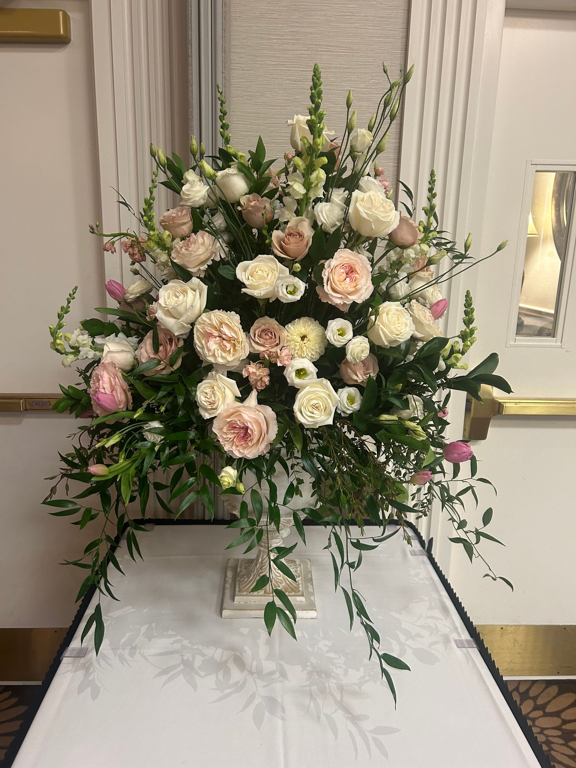 Large floral arrangement of white and blush roses with greenery on a stand in an indoor venue
