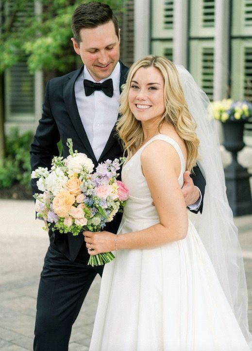 Bride and groom posing outdoors, smiling with a pastel bouquet at a wedding venue