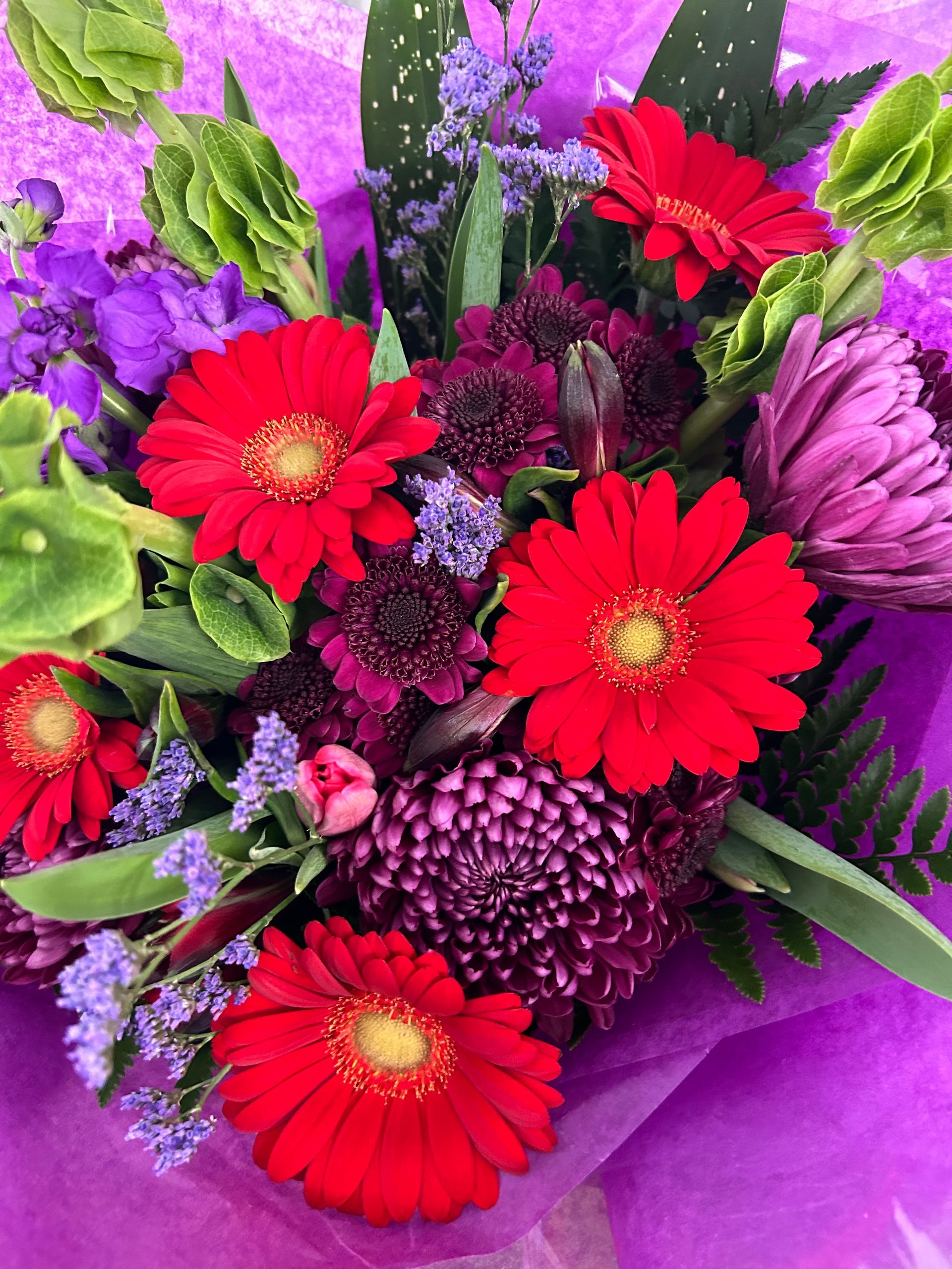 Bouquet of red gerbera daisies, purple flowers, and green foliage wrapped in purple paper