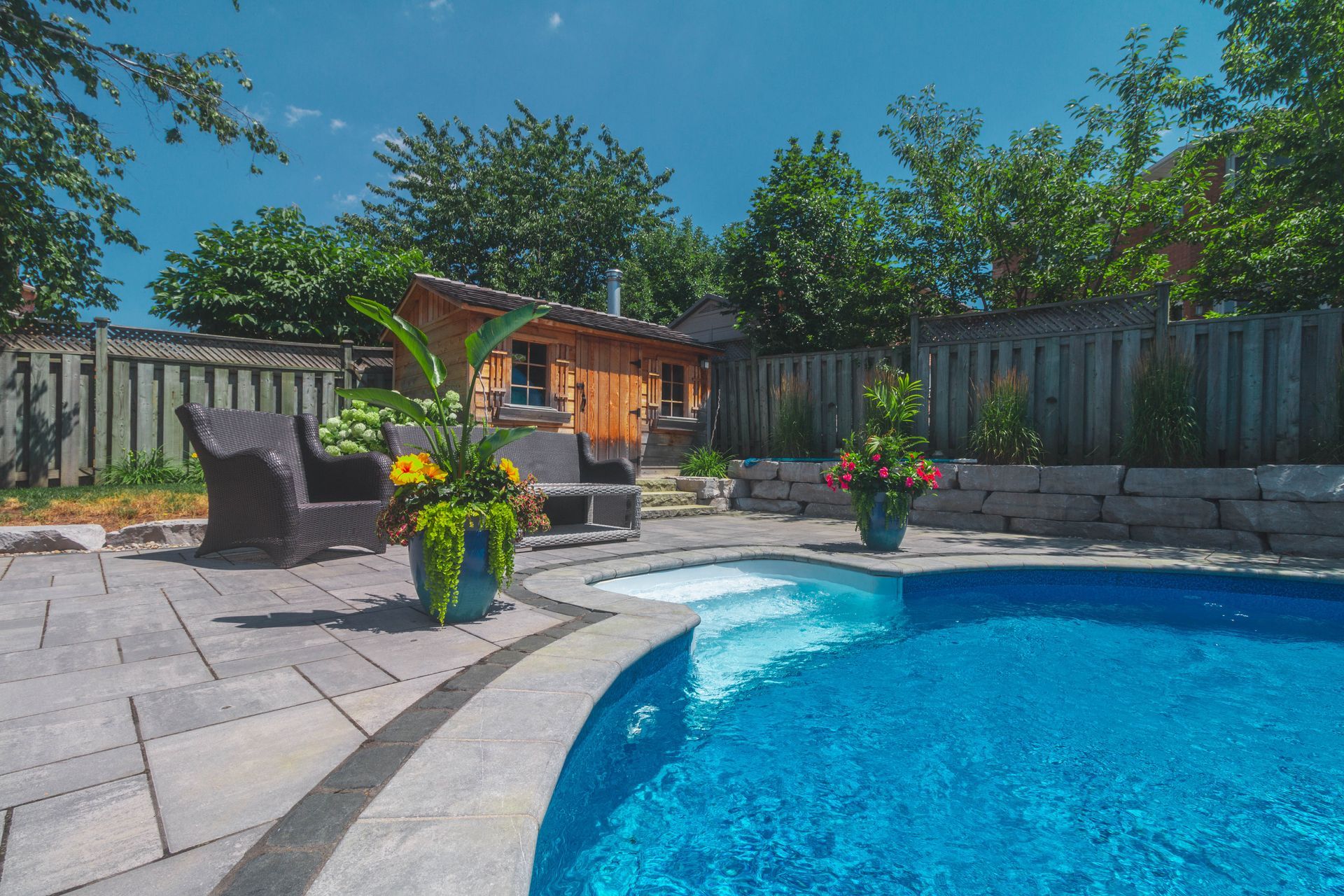 Backyard with a pool, patio, chairs, and small wooden sauna. Sunny day.