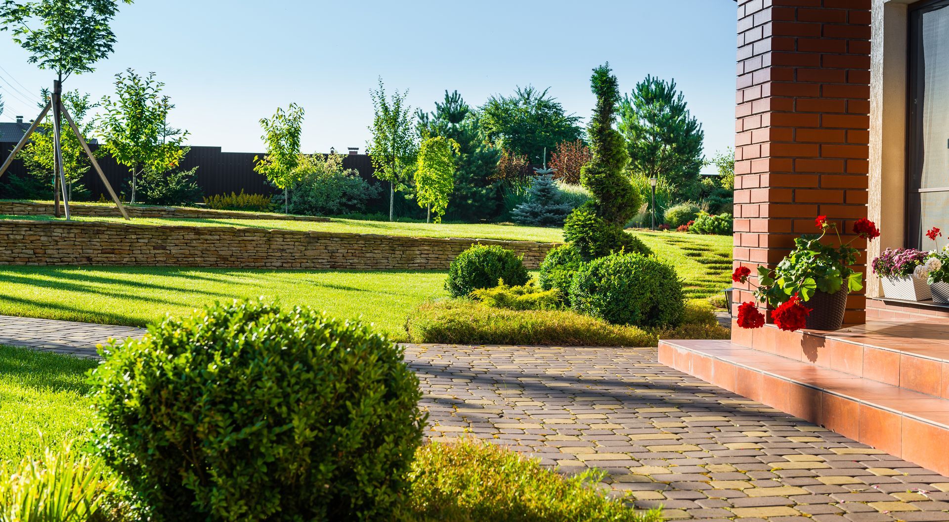 Lush front yard with manicured bushes, brick walkway, and a brick house. Sunny day with green grass and trees.