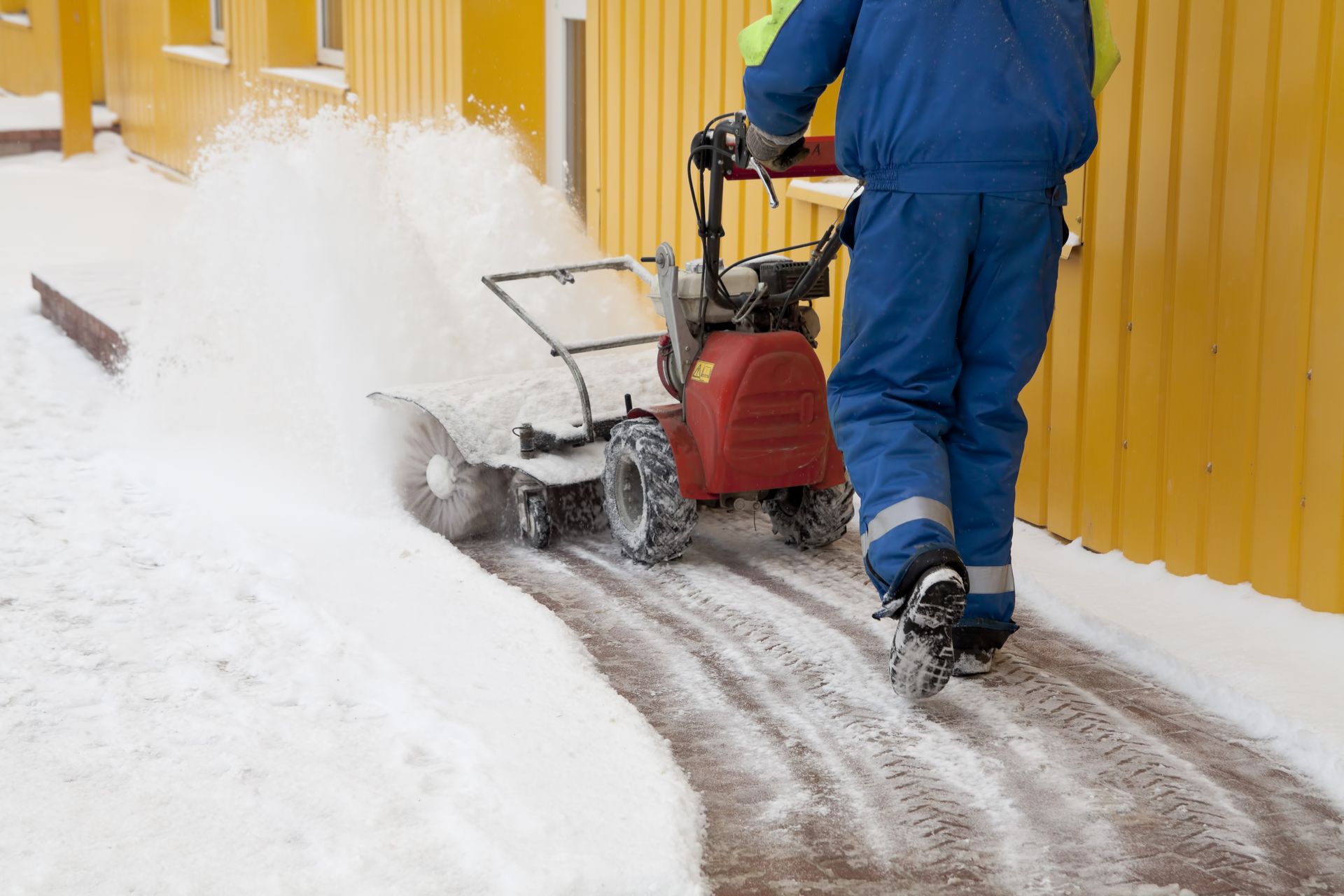 A person in blue overalls uses a snow blower to clear a snowy walkway next to a yellow building.