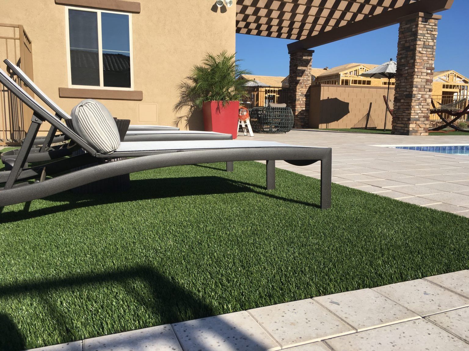 Lounge chairs on green turf beside a pool, near a tan stucco house and pergola.