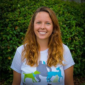 Amy wearing a white t-shirt with dogs on it is smiling in front of a bush.