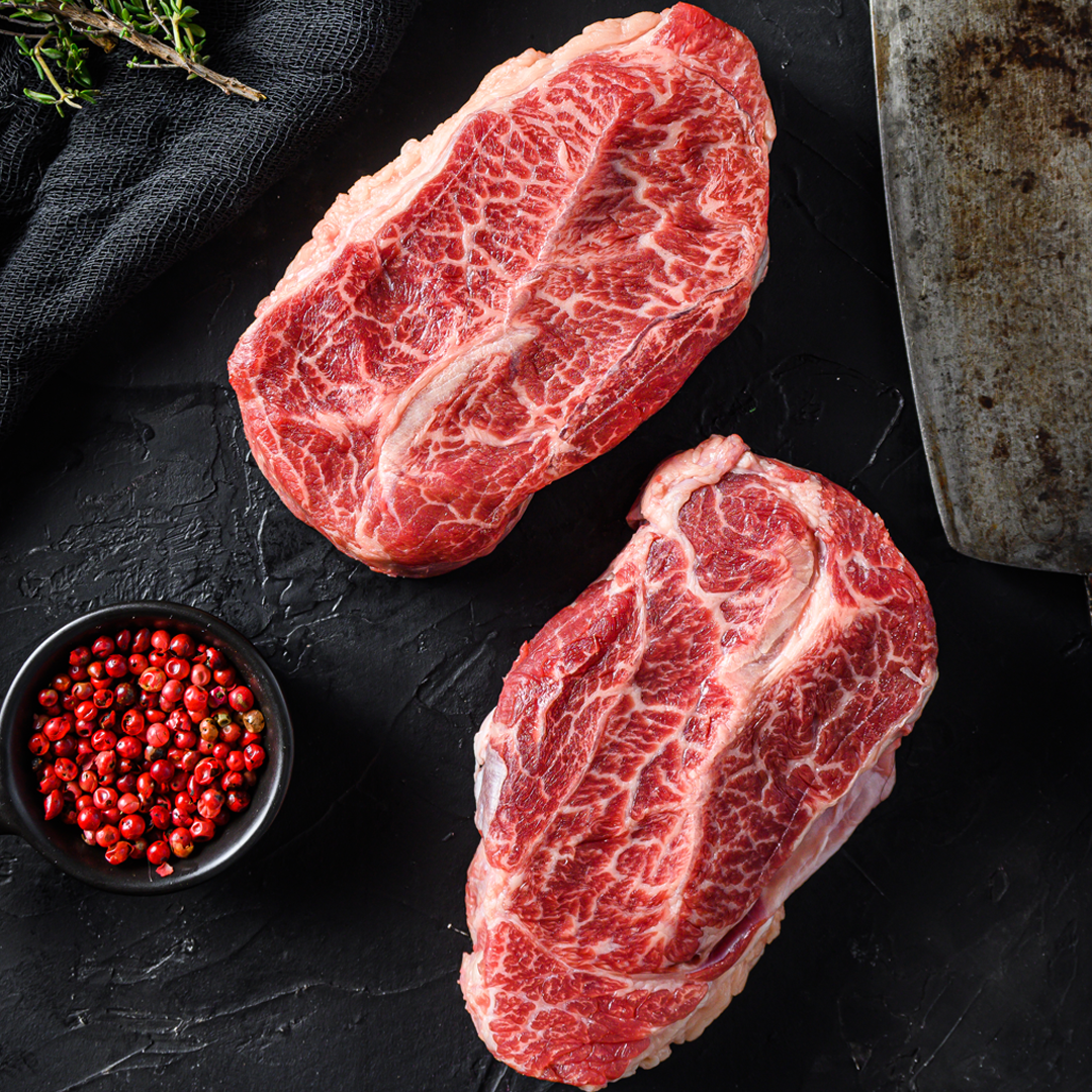 Two raw steaks are sitting on a black table next to a bowl of red pepper.