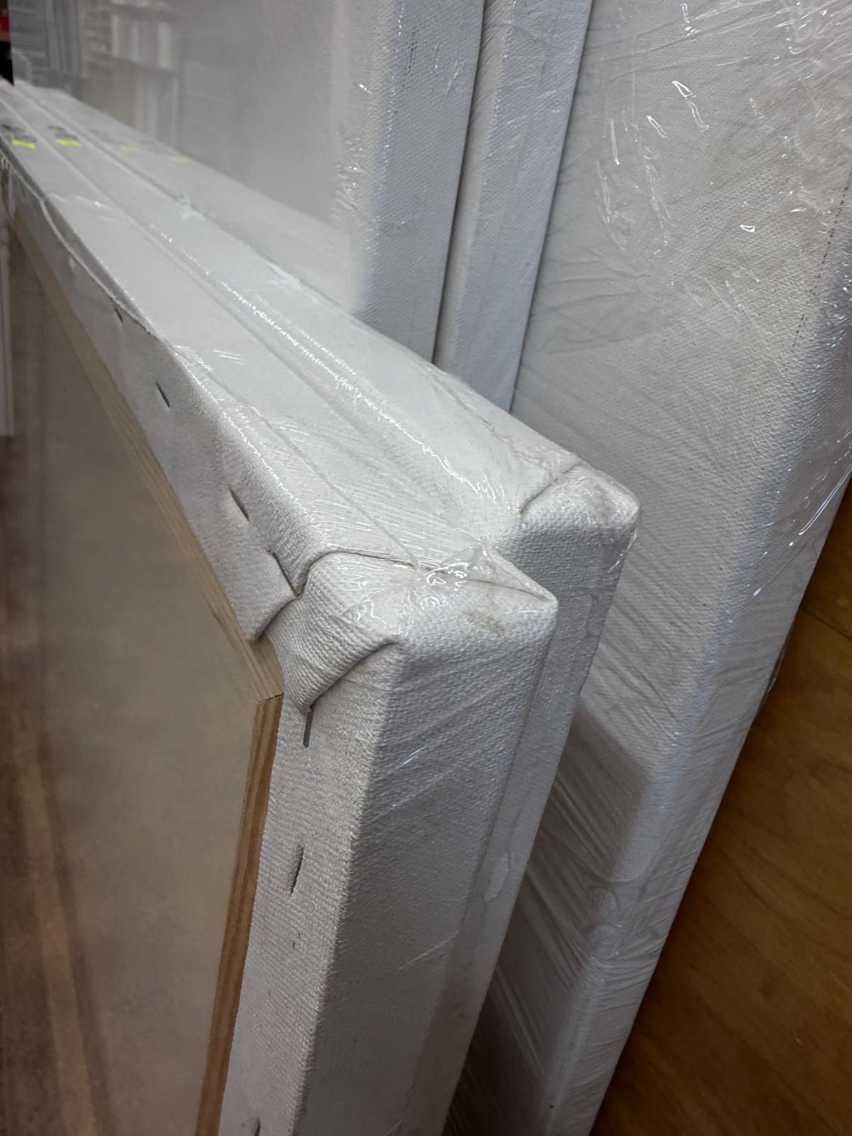 Wrapped white stretcher frames stacked together, with one corner close-up in a warehouse.