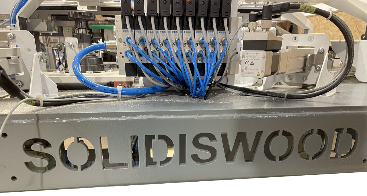 CNC machine with blue hoses and tool heads working on a metal panel labeled “SOLIDISWOOD”