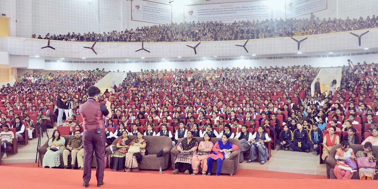 A person stands on a stage addressing a large, seated audience in a packed auditorium with red seating.