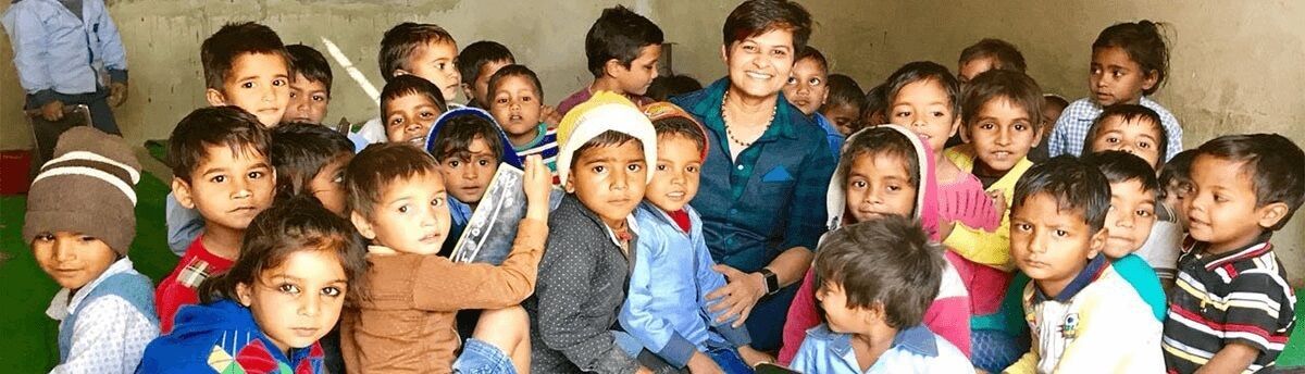 A person smiles while surrounded by a large group of children in a dimly lit, indoor setting.