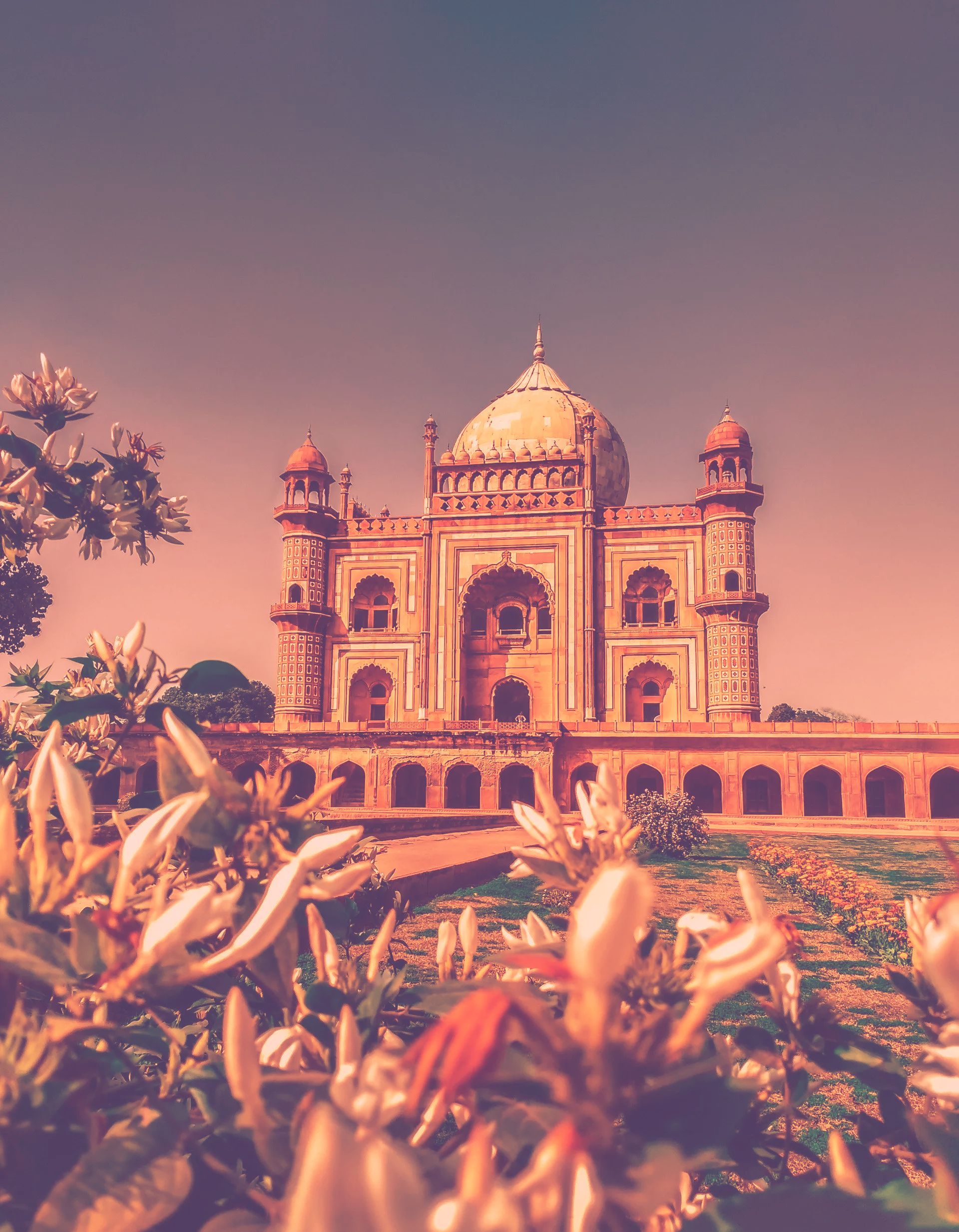 An ornate, symmetrical stone mausoleum with a large dome, framed by out-of-focus white flowers in a warm, sepia-toned light.