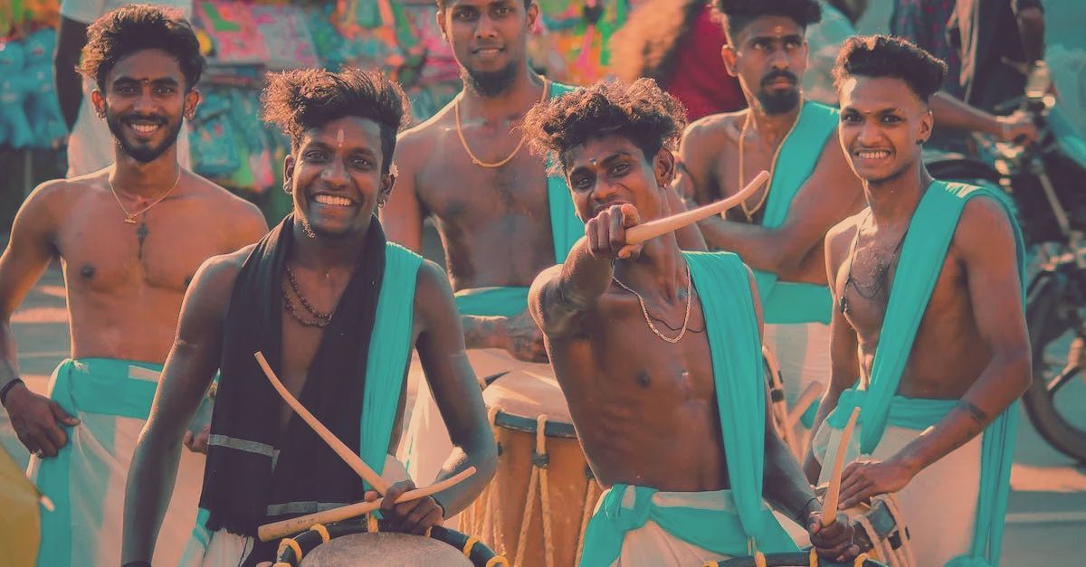 A group of shirtless drummers in teal sashes playing traditional drums outdoors, smiling and gesturing at the camera.