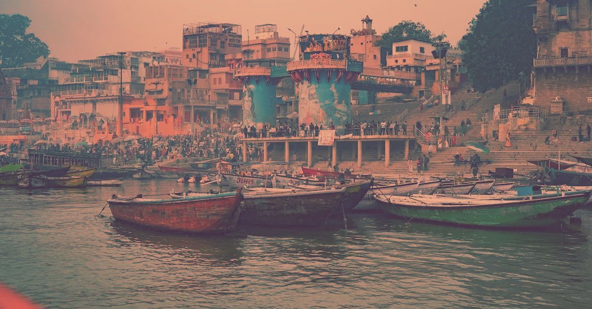 Boats float on the Ganges River in Varanasi, India, before a crowded waterfront of historic buildings and temples.