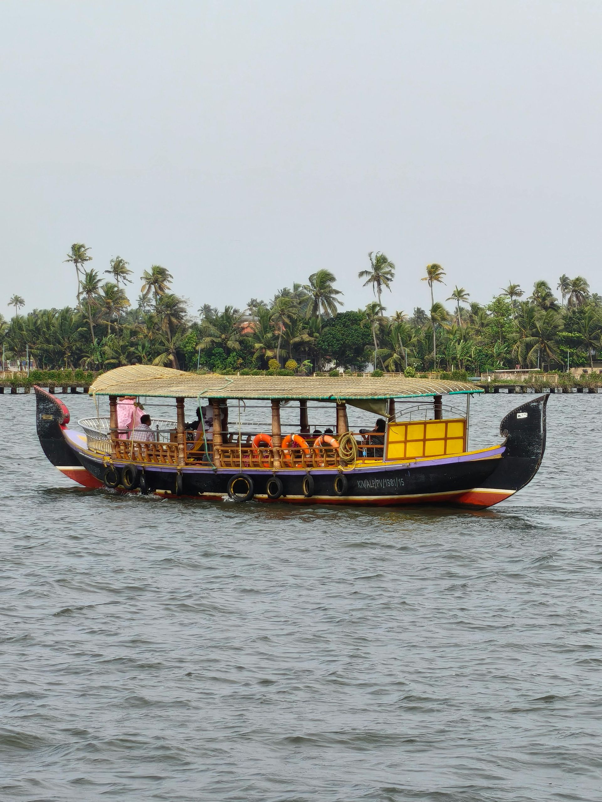 A traditional wooden Kerala houseboat with a thatched roof sails across a calm, open waterway near a palm-lined shore.