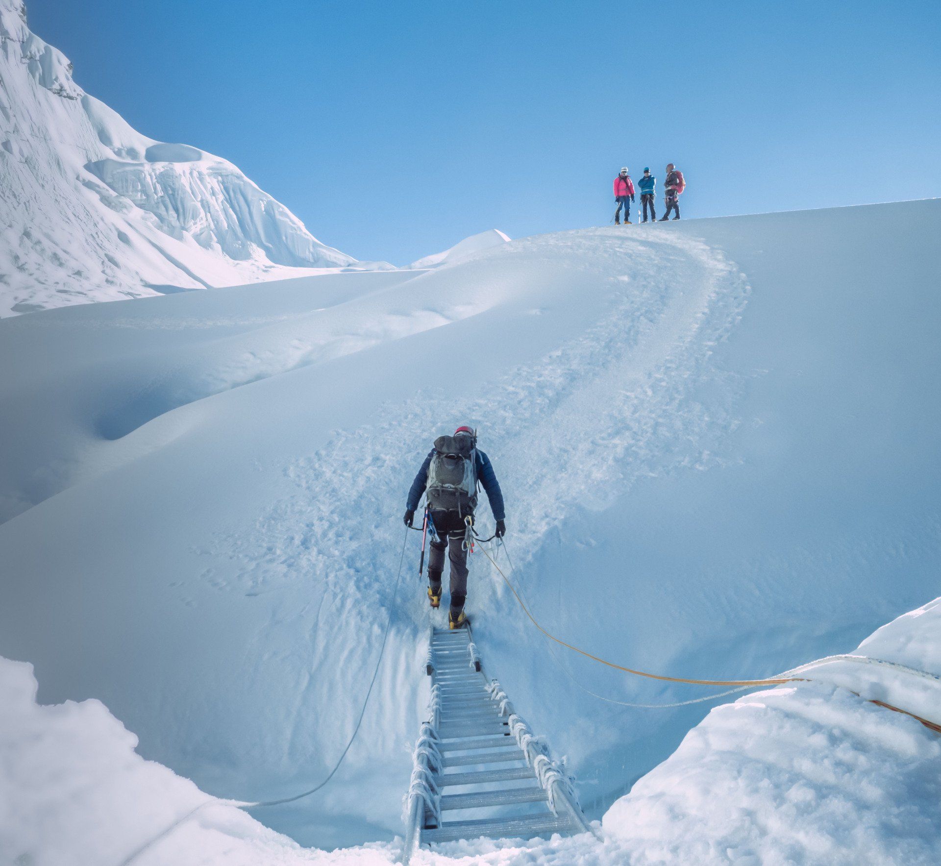 A climber in dark gear carefully traverses a metal ladder over a snowy crevasse, with three others waiting on a ridge.