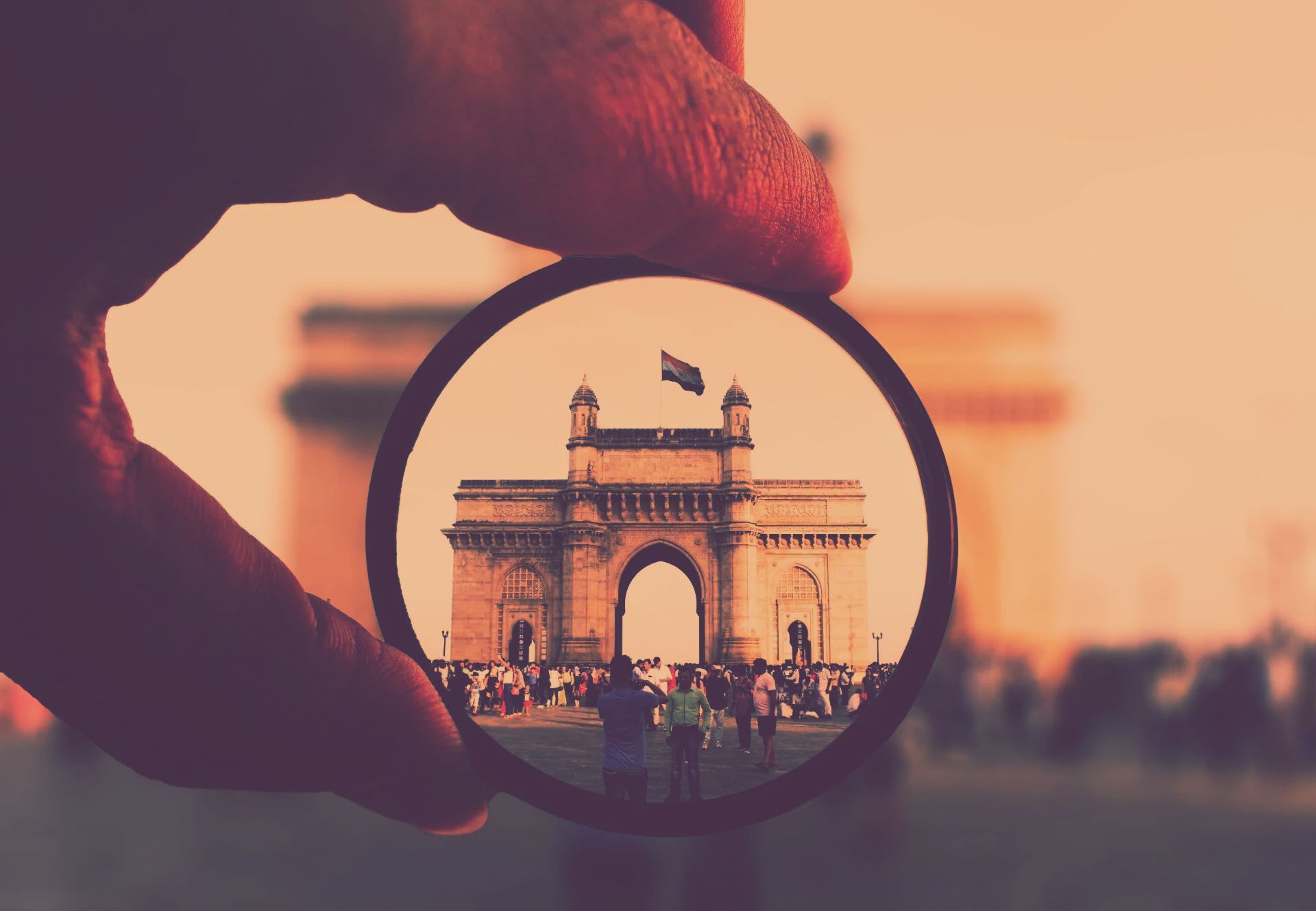 A hand holds a magnifying glass, focusing on the Gateway of India monument with an Indian flag flying on top.