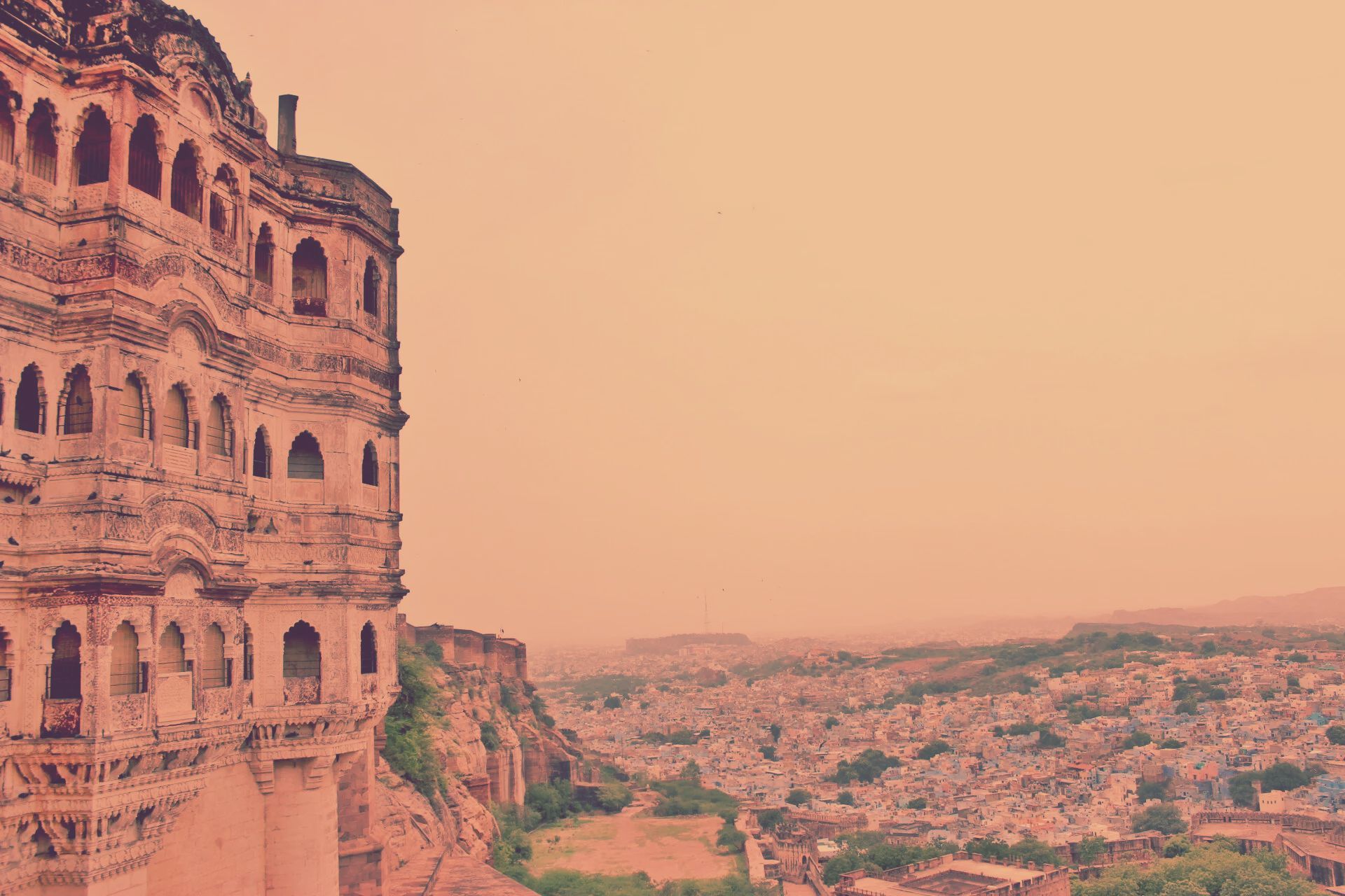 An intricately carved stone palace facade overlooking a sprawling, dense city under a hazy, warm-toned sky.