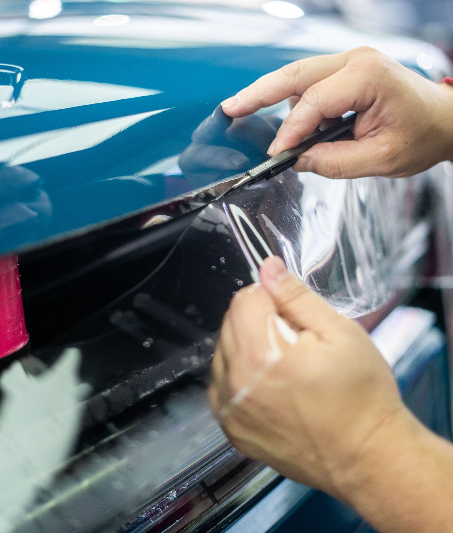Hands applying black trim to a blue car with a tool in an auto repair shop