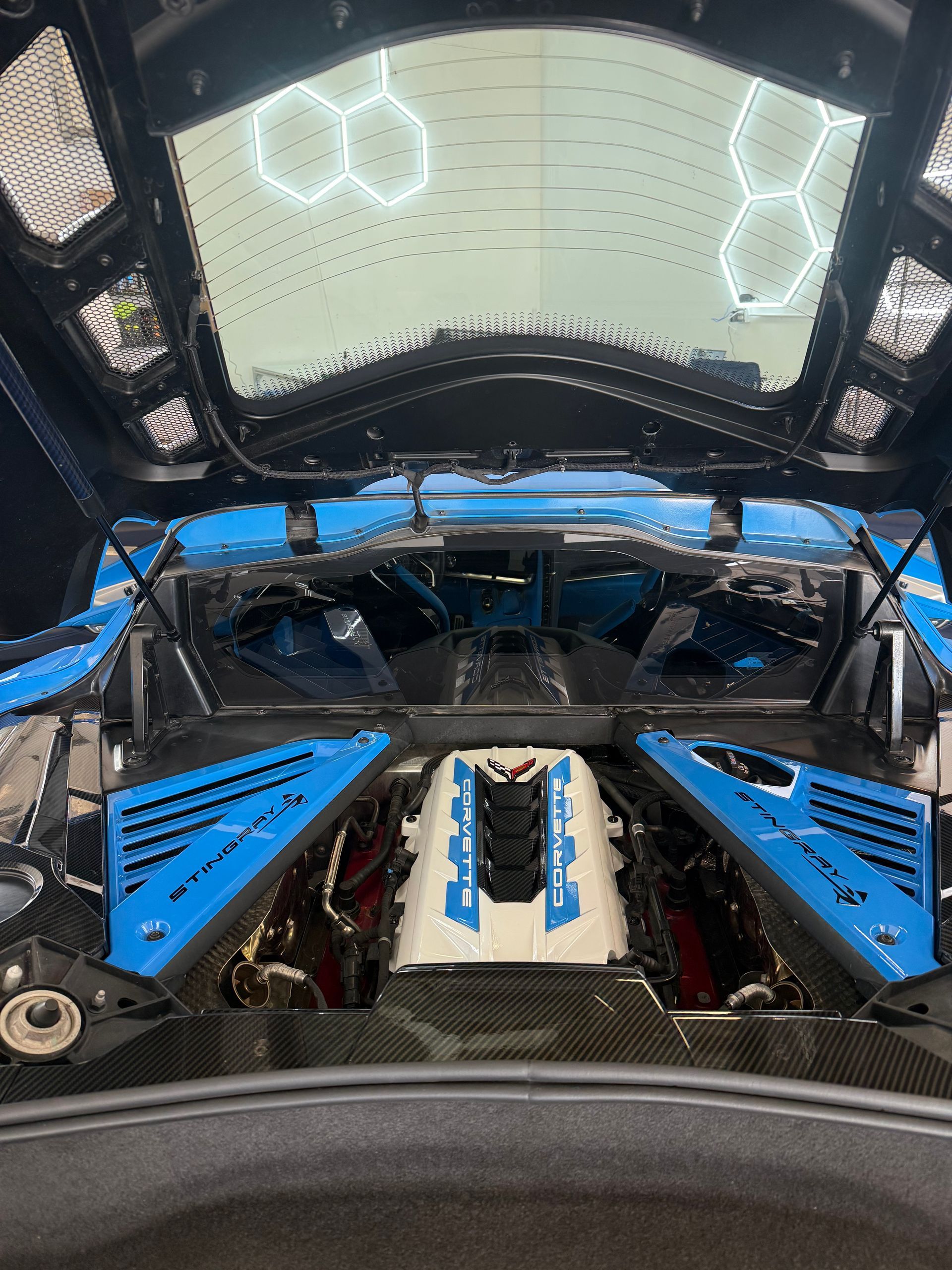 View from inside a blue race car cockpit, showing engine components and a windshield with overhead lights.