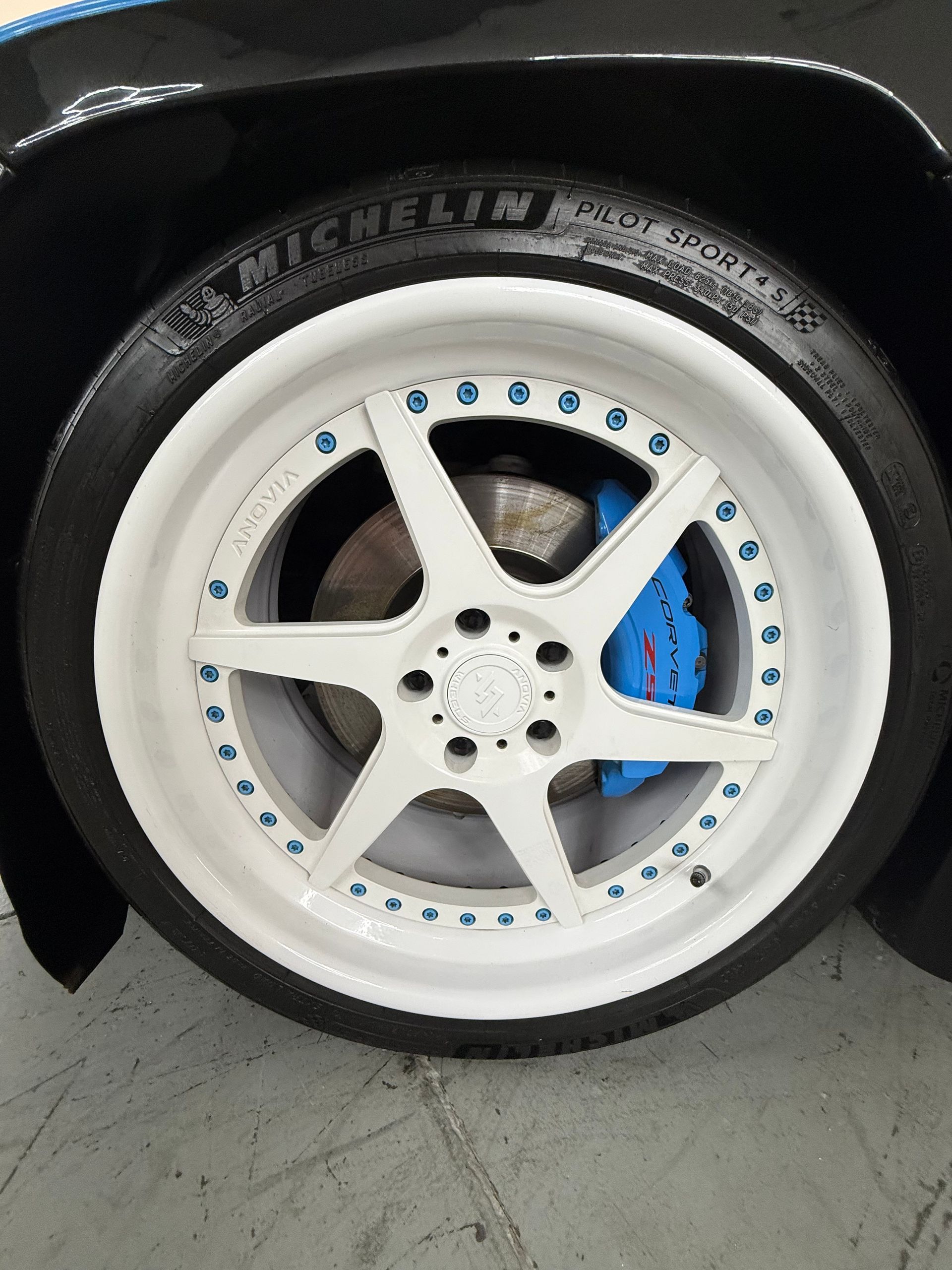 Close-up of a white car wheel with blue brake caliper and black tire on a concrete floor